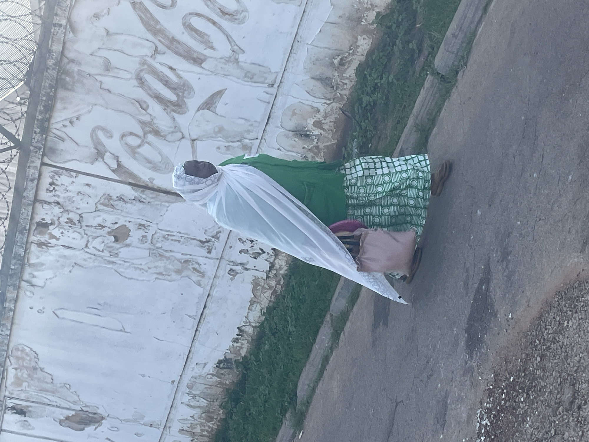 In Jos, Nigeria, a woman walks along a cracked asphalt road next to a grassy embankment. She wears a vibrant green dress with a white and green geometric pattern, a white headscarf, and brown sandals. A light pink shoulder bag is slung over her left arm. Behind her, a distressed white wall with peeling paint is visible, and a faded Coca-Cola logo is partially discernible. Barbed wire runs along the top of the wall, suggesting a security perimeter. The lighting indicates it is daytime, with the sun casting shadows.