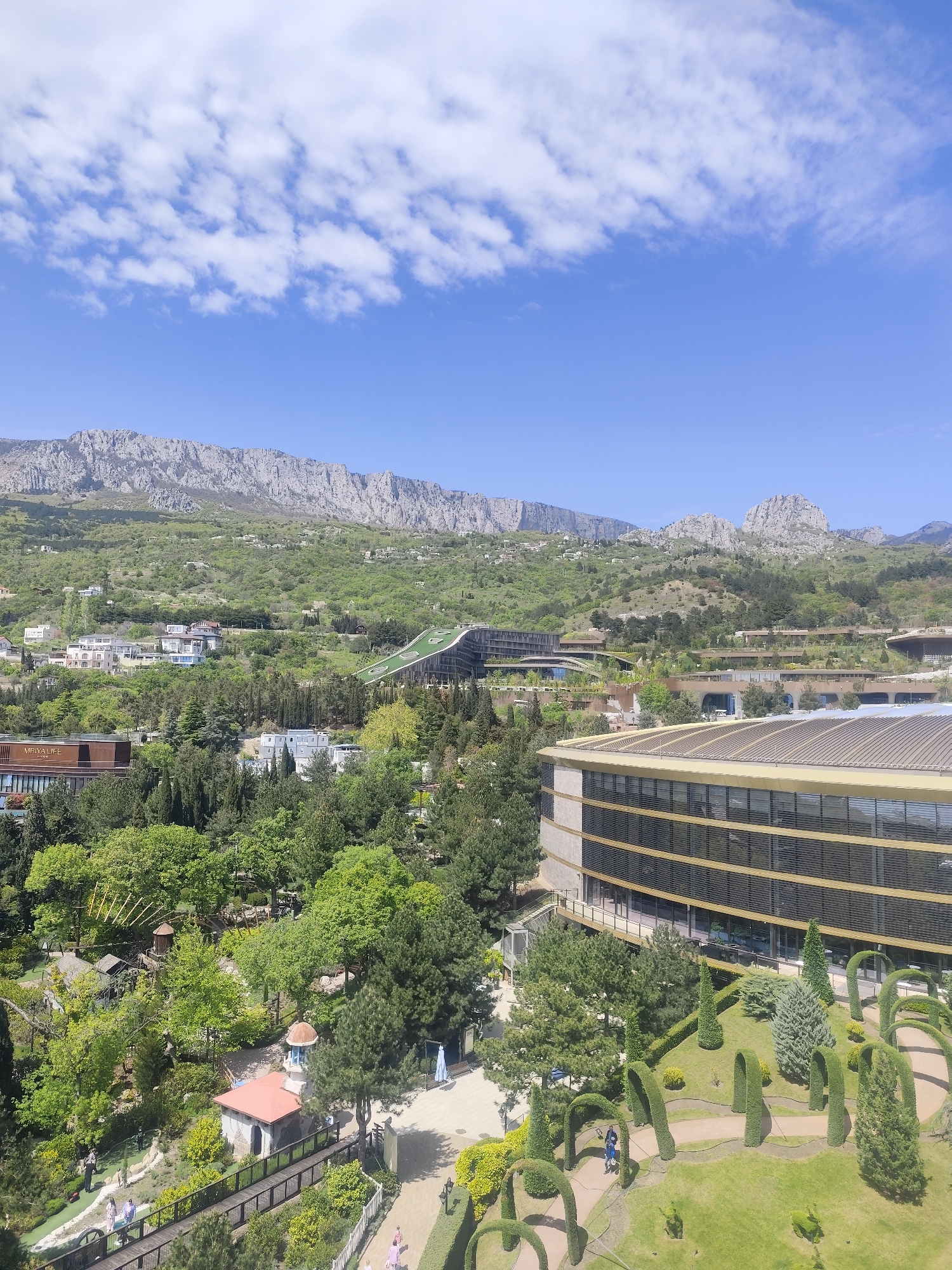 The image shows a scenic view in Симеизский поселковый совет, Ukraine, with a mountainous backdrop under a clear blue sky with scattered clouds. In the foreground, there is a landscaped park with geometrically trimmed bushes and trees, and walkways with people strolling through. To the right, a modern building with large windows dominates the scene. Beyond the park, there are more buildings nestled among green hillsides leading up to the mountains in the distance.