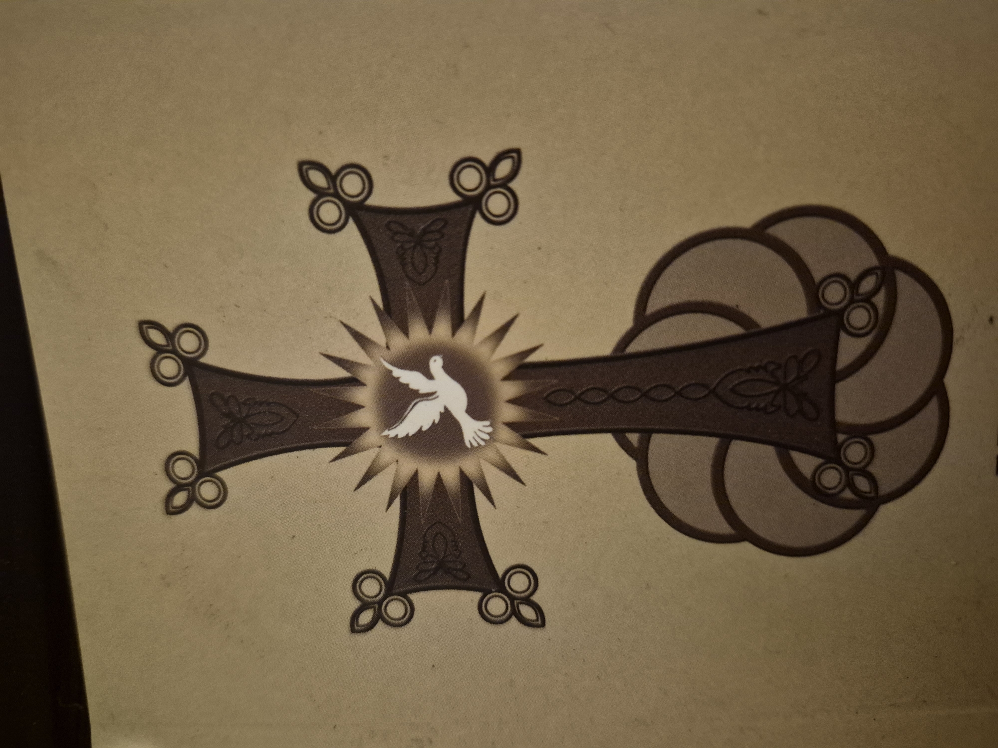The image depicts a stylized Christian cross, likely an Armenian cross ("Khachkar"), with a white dove superimposed at the center. The cross is brown with decorative elements on each arm, including stylized trefoil shapes. The dove is centered in a burst of light or sun rays. The background is a beige or tan color, possibly paper or fabric with slight variations in tone. There is no information in the image to provide location in the city of Yerevan, Armenia.