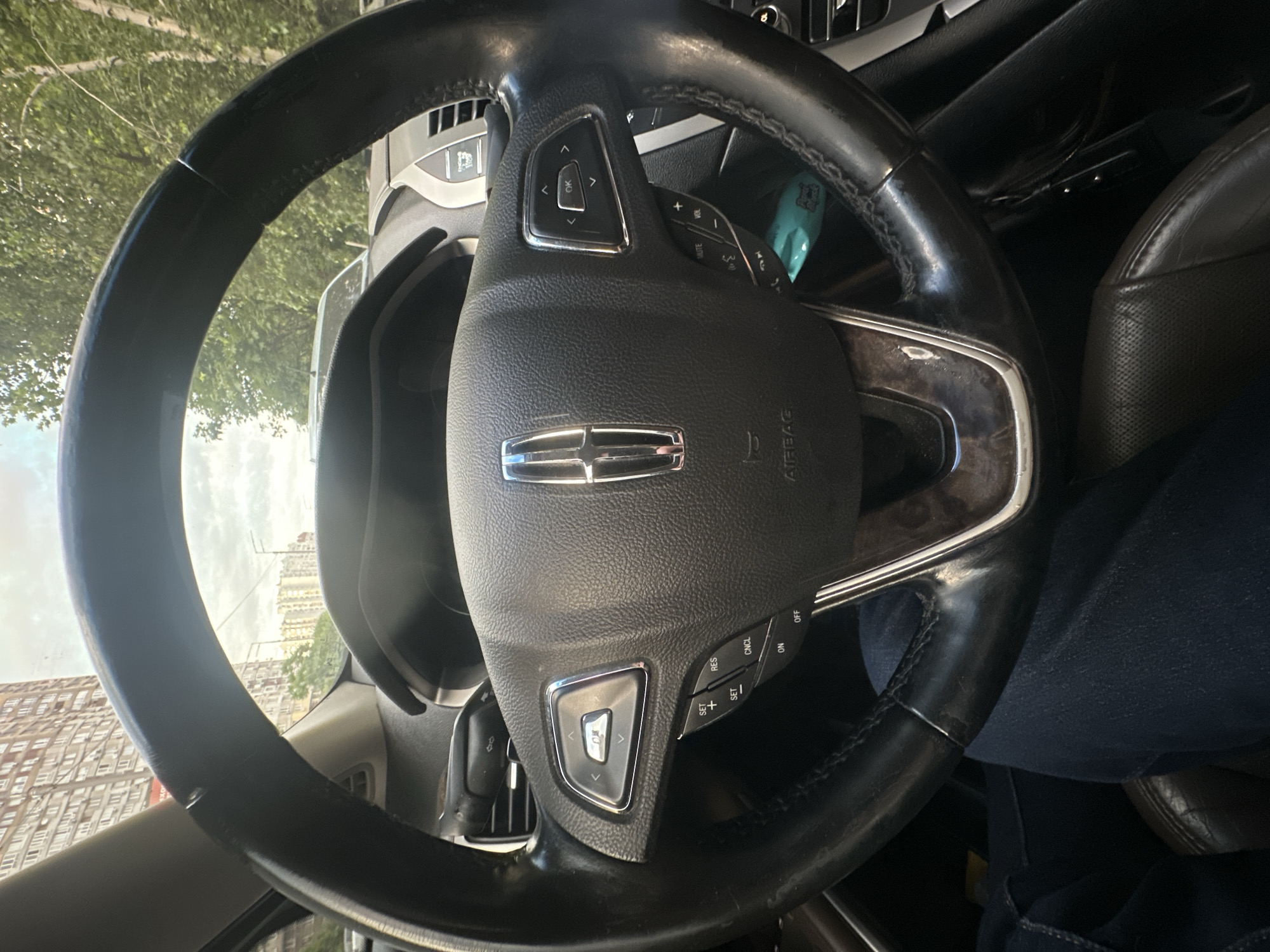 The image is a first-person view from inside a car, looking at the steering wheel and dashboard. The steering wheel is black leather with silver accents and controls on the left and right sides. A Lincoln logo is prominently displayed in the center of the steering wheel. The dashboard features various buttons and a section with wood grain trim. Outside the car, through the windshield, tall apartment buildings and trees are visible, suggesting an urban environment. The sky is cloudy. The car appears to be stationary in what is likely a street or parking area in Kyiv, Ukraine, based on the architectural style of the buildings. A person's legs, clad in dark blue jeans, are visible in the lower part of the frame, indicating the driver or a passenger.