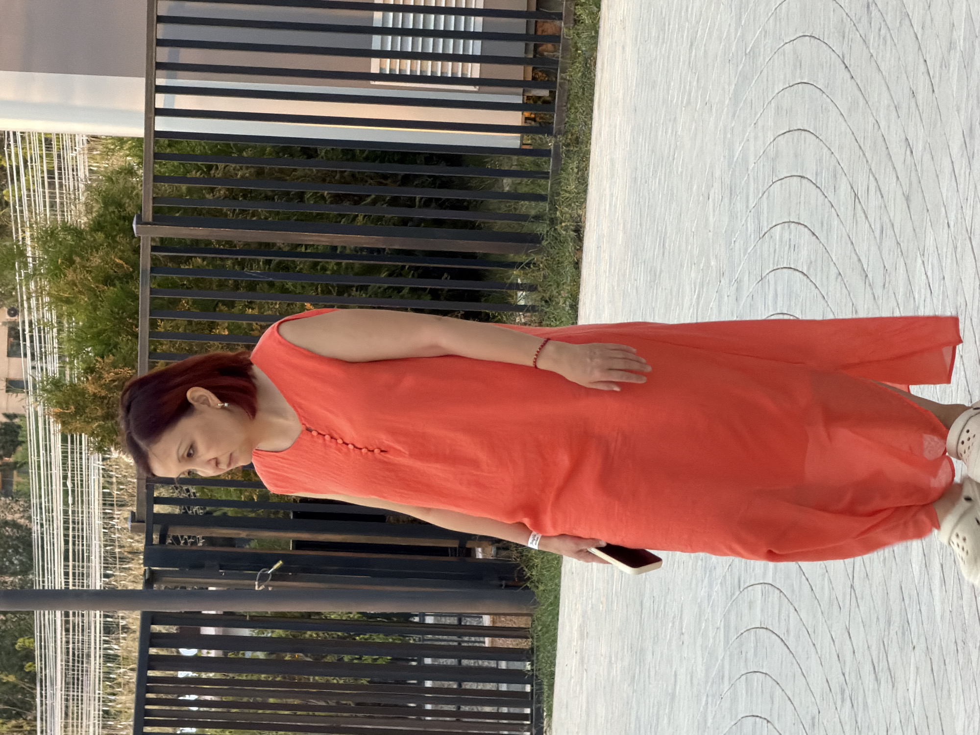 A woman with short, dark red hair is walking outdoors. She is wearing a bright orange, sleeveless dress that reaches her ankles and white clogs. The woman is holding a smartphone in her right hand. She is wearing a white wristband on her left wrist and a red beaded bracelet on her right wrist. She is looking to her left. Behind her is a dark metal fence, green bushes, and a building with white vertical elements and horizontal vents. The ground is paved with light-colored stones in a circular pattern. The scene appears to be in a public area.