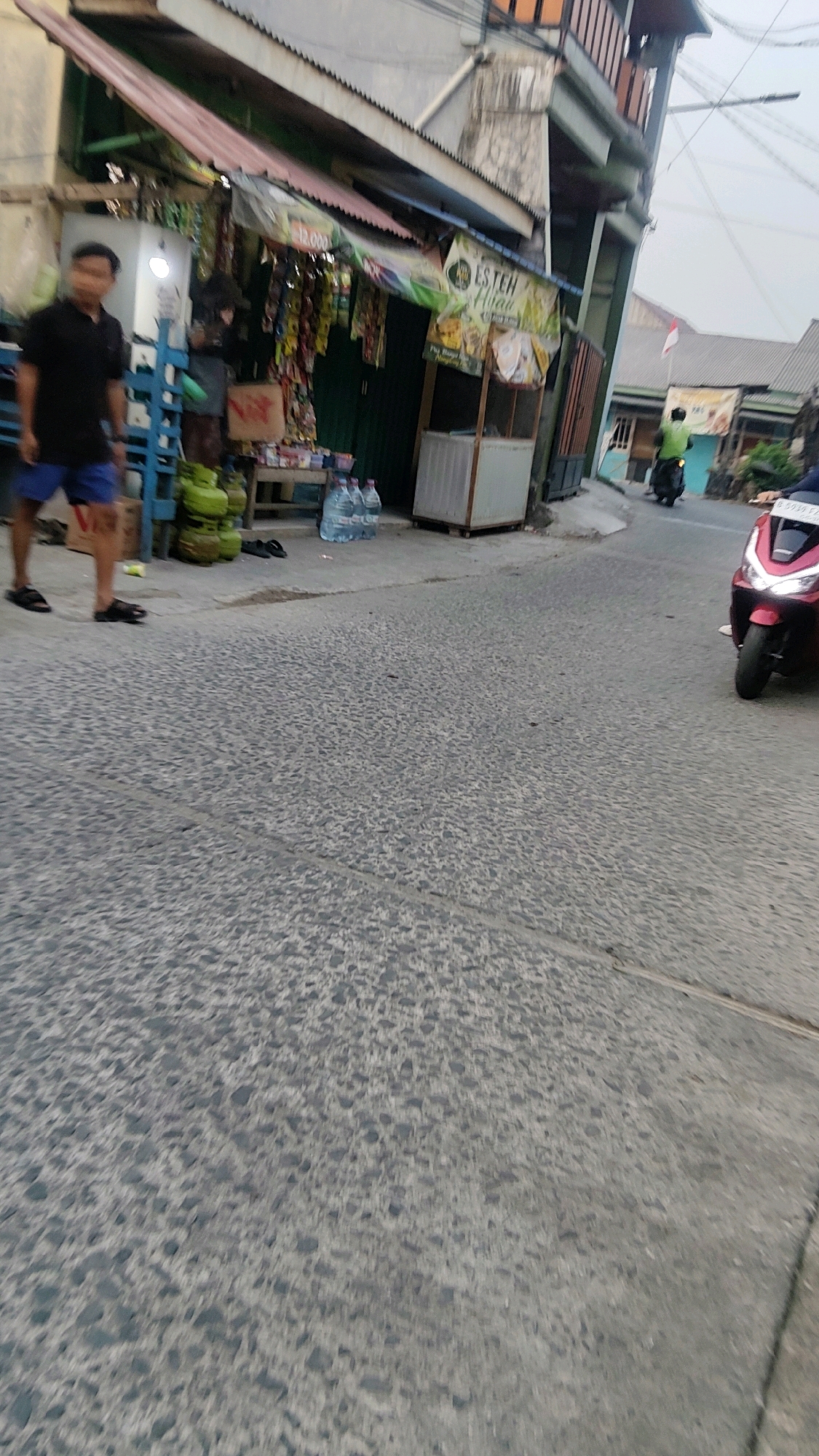 In the city of Mekarmukti, Indonesia, a street scene unfolds with a small shop on the left side of the road. The shop features a red and pink awning with various items for sale displayed both inside and outside, including what appear to be snacks and drinks. Several green gas cylinders are stacked near the shop. A young man wearing a black t-shirt and blue shorts stands outside the shop, facing forward. Further down the street, a person is riding a red motorcycle, and another person is on a dark-colored motorcycle further in the distance. The road surface is asphalt with a textured pattern. The overall environment suggests a typical street in a residential or commercial area.