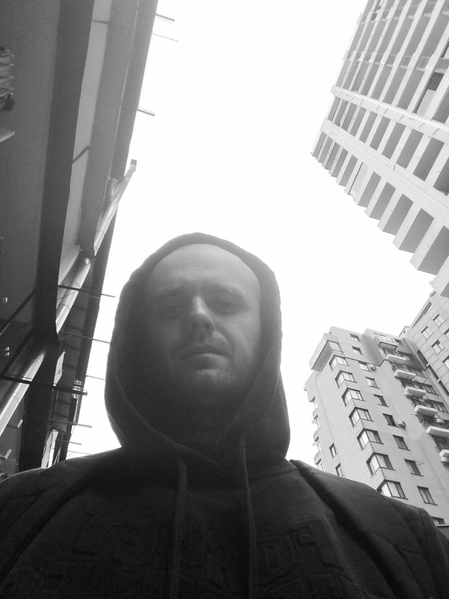 A black and white, low-angle, selfie-style photograph shows a man wearing a hooded sweatshirt. The man's face is centered in the frame, and he appears to be looking directly at the camera. His hood is pulled up, obscuring the top of his head. The background consists of tall, modern buildings, characteristic of a city environment. One building on the left appears to be a more industrial or older structure with visible pipes and scaffolding, while the buildings on the right are contemporary residential or office towers with repeating patterns of windows and balconies. The sky is overcast and bright, suggesting daytime. The composition creates a dramatic perspective, emphasizing the scale of the buildings in relation to the man. The overall impression is urban and slightly gritty, with the man at the forefront against a backdrop of towering architecture.