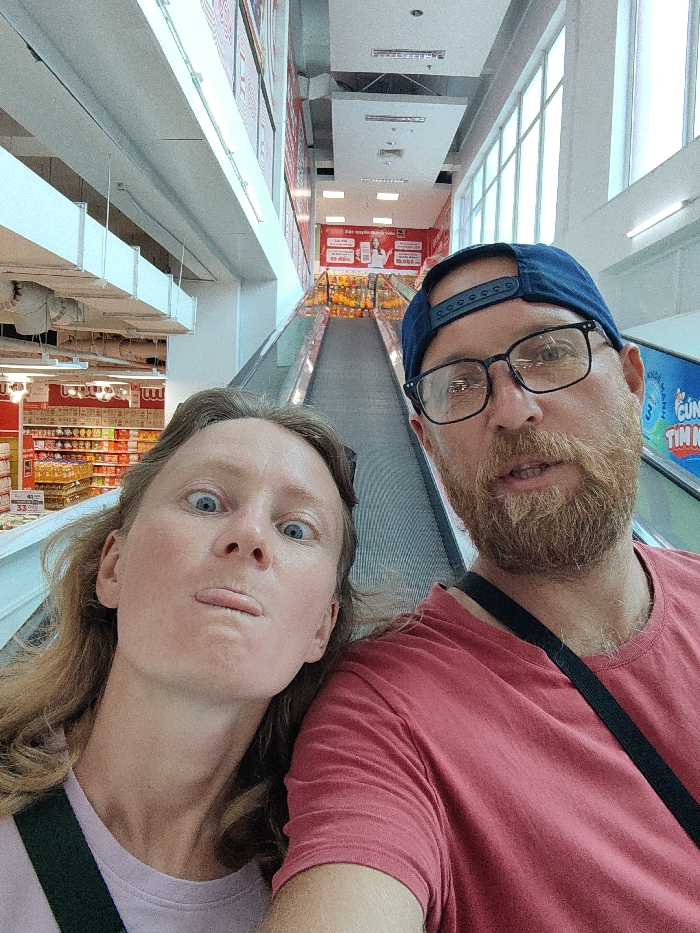 The image captures two individuals on an upward-moving escalator within a large retail establishment in Thuận Hóa Ward, Vietnam. The scene is brightly lit, indicative of a supermarket or department store interior.
In the foreground, an adult female on the left has light skin, light brown hair, and blue eyes. Her eyes are wide open, and she exhibits a playful facial expression with her tongue slightly extended between closed lips. She wears a light pink or purple shirt, with a dark strap visible over her left shoulder. To her right, an adult male with light skin, a reddish-brown beard, and blue eyes wears black-rimmed glasses and a dark blue baseball cap reversed. He is dressed in a reddish-pink t-shirt, and a dark strap is visible across his right shoulder. His mouth is slightly open. Both individuals are looking directly towards the camera.
The background reveals the commercial environment. On the left side, multi-tiered retail shelving is stocked with various products, including numerous bottles with yellow contents and other packaged goods, under red and white signs. On the right, alongside the escalator, large red and white promotional banners with Vietnamese text and a QR code are affixed to the wall. Above, ceiling structures feature strip lighting. Large multi-paned windows are visible on the upper right, and another escalator ascends in the deeper background. The flooring and additional shelving are visible beyond the escalators.