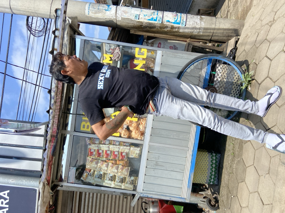 A man, wearing a black t-shirt with the words "SEXY" and "0284" printed on it, light blue jeans, and white flip-flops, is standing to the right of a food cart in the city of Padalarang, Indonesia. He is facing to the right and looking in that direction. The cart is silver and blue, with a glass front that displays snacks and drinks. There is a blue wheel on the right side of the cart. The man is leaning to the left side, and his right hand is resting on his hip. The background is a busy street with buildings, wires, and a blue sky. The location of the scene is identified by the provided coordinates, which correspond to a street in Padalarang, Indonesia.
