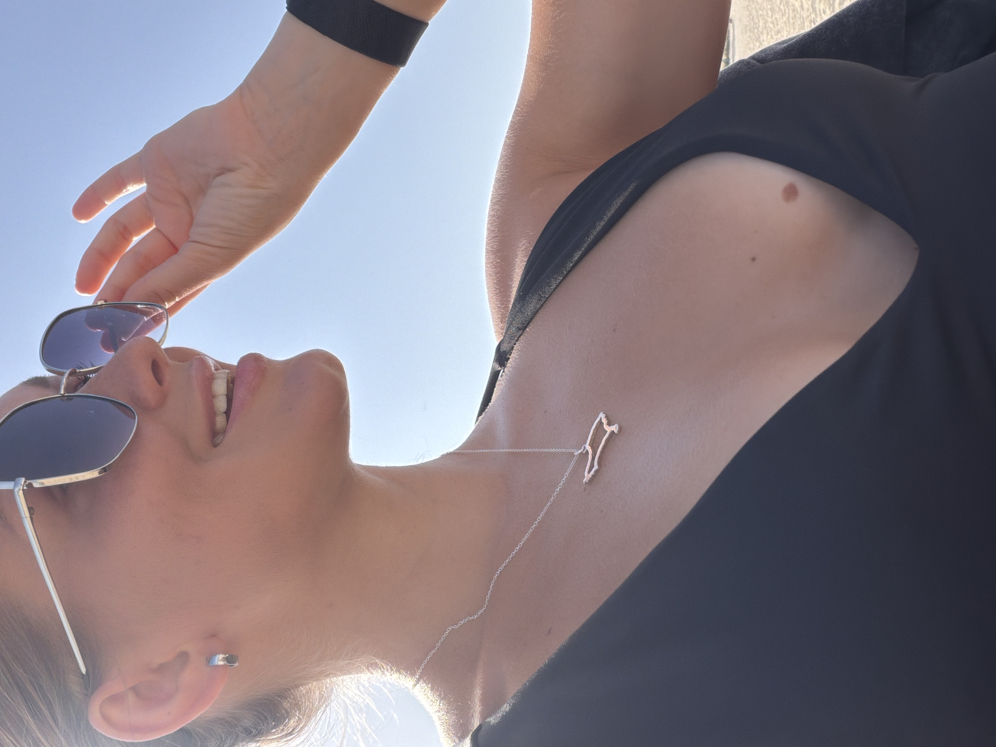 A low-angle, close-up shot shows a woman smiling and adjusting her sunglasses on a sunny day. She wears a black top, a silver chain necklace with a pendant in the shape of a country, and a black wristband on her arm. There is a mole on her chest and a small earring in her ear. The background is a clear, bright sky. There is no evidence to confirm this photo was taken in Dubai, United Arab Emirates.