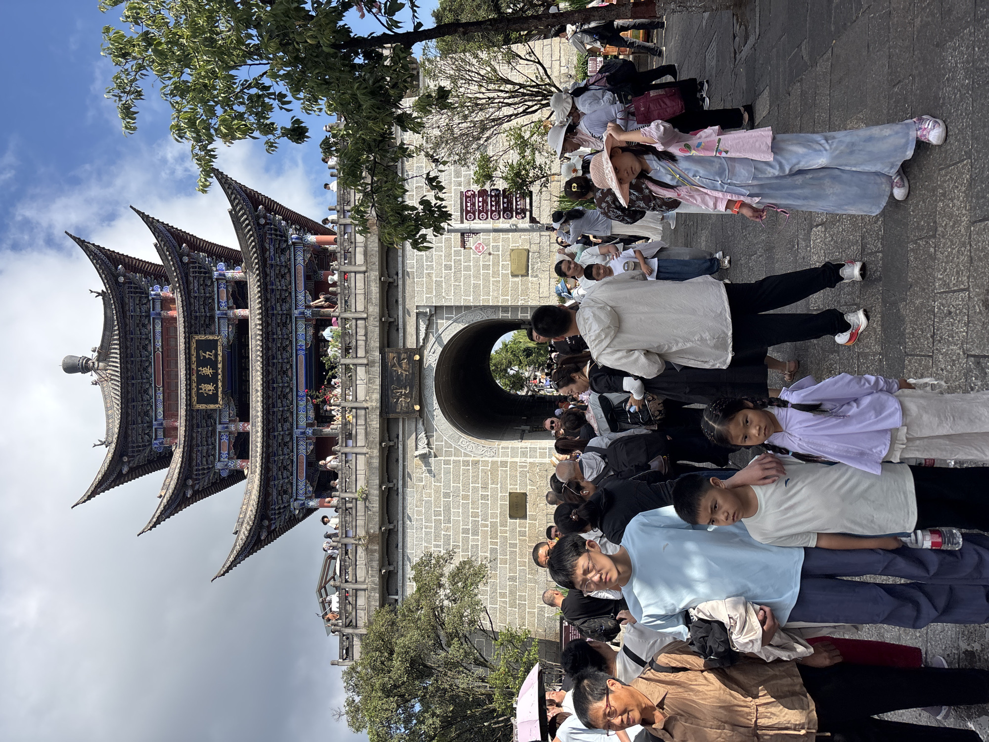 The image captures a lively scene at the gate of Dali City, China. A traditional multi-tiered pagoda structure stands prominently above a stone archway, with people gathered on a balcony overlooking the bustling street below. The area is filled with a diverse crowd, including families, tourists, and locals, dressed in a mix of casual and traditional attire. Many are seen walking, talking, and taking in the sights. A young girl in a pink hat and blue outfit stands out in the foreground, looking towards the camera. The ground is paved with cobblestones, and lush green trees add to the vibrant atmosphere. The sky is partly cloudy, suggesting a pleasant day for exploration.