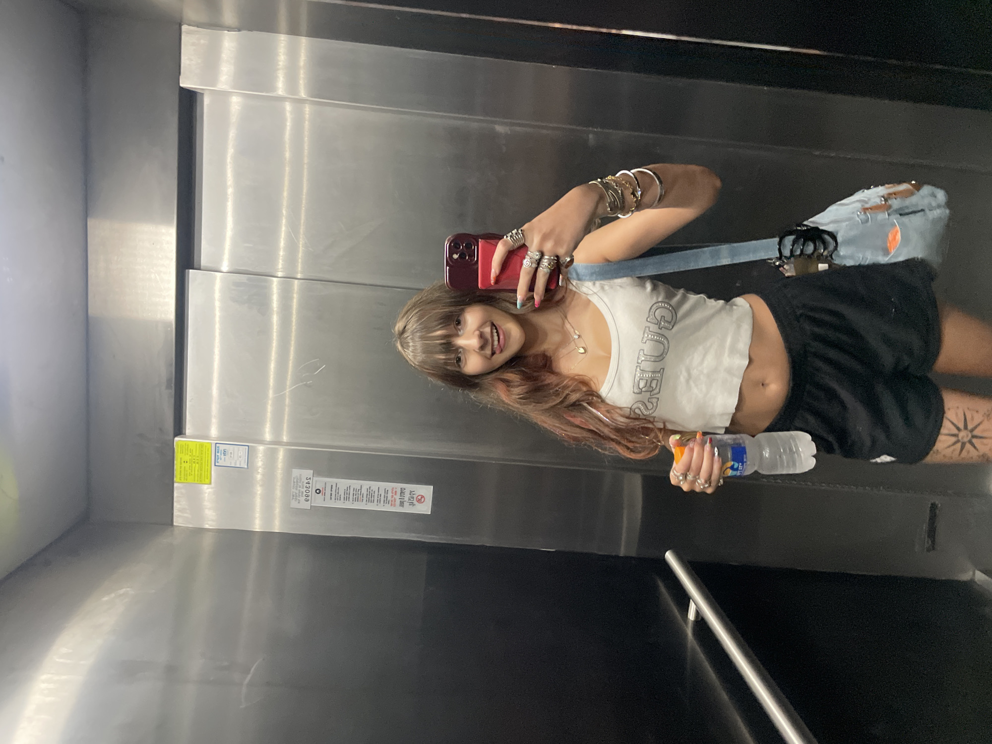 A young woman with bangs and reddish-brown hair takes a selfie in an elevator. She is wearing a white crop top with "Guess" written on it, black shorts, and numerous rings on her fingers. She is holding a bottle of water in her right hand and a phone in her left. A light blue distressed denim bag hangs from her shoulder. The elevator is made of stainless steel, with a safety bar on the left side and various labels on the right wall. The image appears to have been taken in Raanana, Israel, as indicated by the labels on the elevator wall which include Hebrew text and what appears to be a city sticker from Raanana.