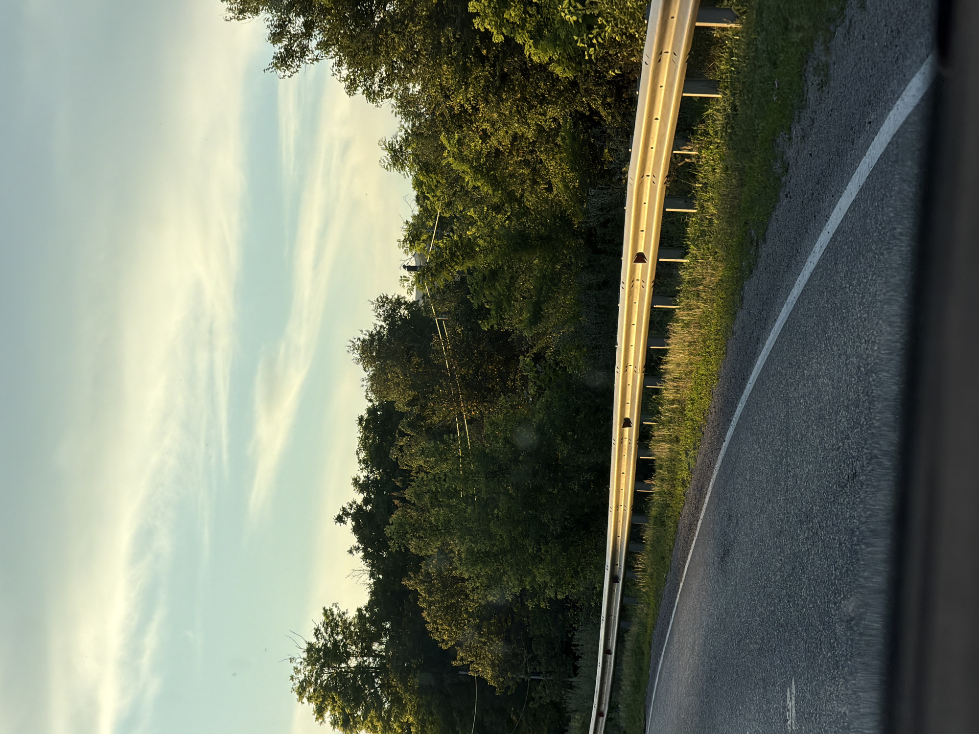 This media file shows a roadside view from a vehicle in Aleksandrovskoye, Russia. The scene depicts a paved road curving to the right, bordered by a yellow guardrail on the left. Lush green trees and foliage line the other side of the guardrail, with some of the trees appearing quite tall and dense. The sky is a pale blue with wispy white clouds, suggesting it is daytime, possibly late afternoon or early evening given the soft lighting and shadows. The overall atmosphere is peaceful and natural, with no people or specific events visible. The image captures a typical rural or semi-rural roadside environment.