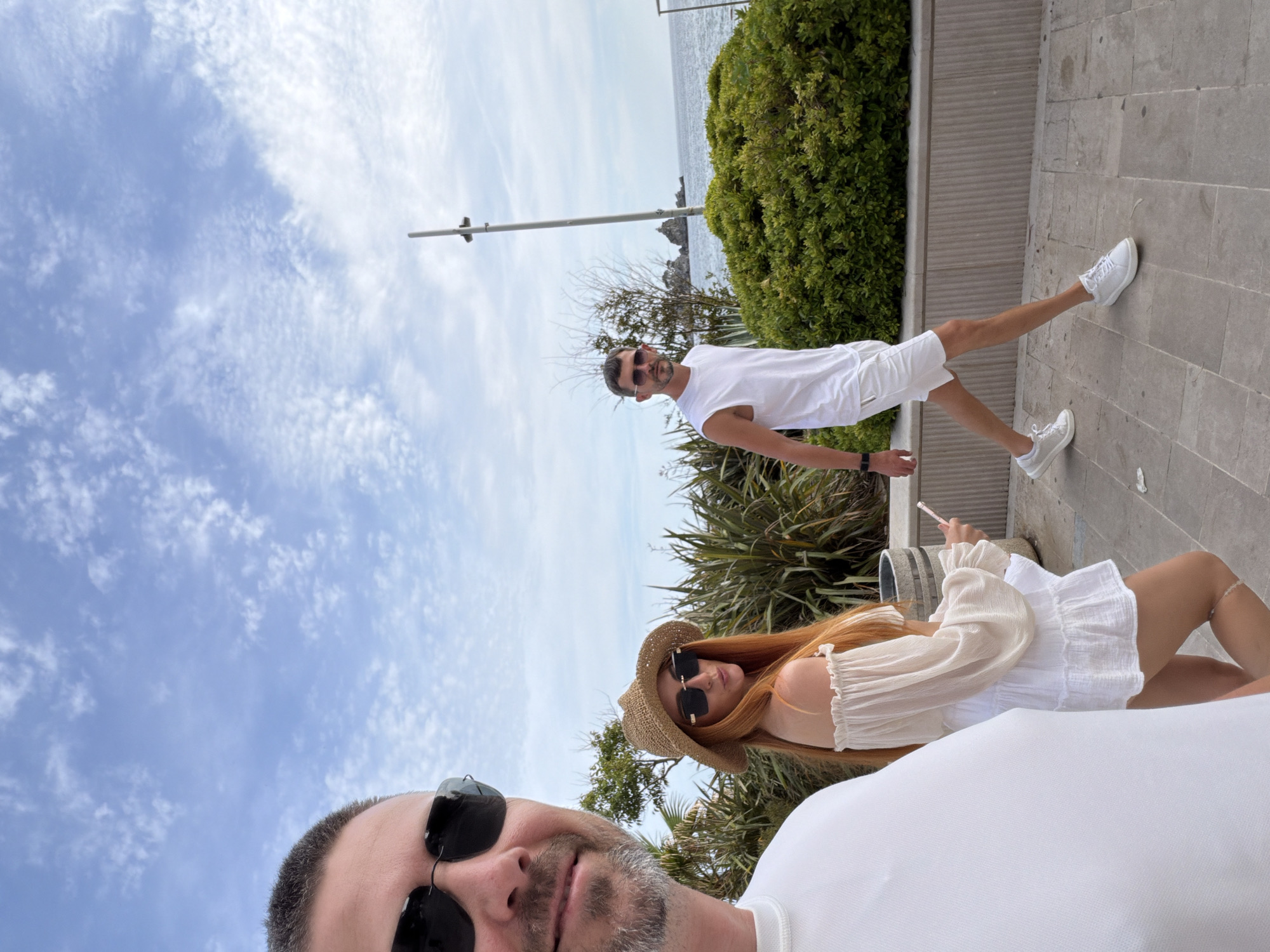 The image depicts three individuals on a paved walkway in Petrovac centar, Montenegro, adjacent to the sea under a partly cloudy sky.
In the immediate foreground on the left, a male subject is partially visible from the forehead to the upper chest. He wears dark sunglasses and a white crew-neck shirt. His hair is short and dark, and he has light stubble or a short beard. He is facing slightly towards the viewer, suggesting he is taking the photograph or is closest to the camera.
In the mid-ground, slightly to the left of center, a female subject walks to the right. She has long, bright reddish-orange hair and wears a straw hat and dark, square-framed sunglasses. Her attire consists of an off-the-shoulder, long-sleeved, light-colored (possibly beige or off-white) top and a short, tiered white skirt. A white anklet is visible on her right ankle. She holds a small pink object in her right hand and is looking back towards the camera over her left shoulder. Behind her, a round, metallic waste bin is partially visible.
Further in the mid-ground, to the right, a second male subject walks to the right, slightly ahead of the female. He wears dark sunglasses, a white sleeveless top, white shorts, and white sneakers. A dark watch is on his left wrist. He has short, dark hair and a short beard, and is looking towards the camera.
The walkway is paved with light-colored rectangular flagstones. A low, fluted concrete retaining wall separates the walkway from a planted area containing dense green bushes and spiky plants. Beyond this vegetation, the Adriatic Sea is visible with rocky outcrops in the distance. A tall, light-colored utility or light pole stands upright in the background on the right. The sky is light blue with scattered white, cumulus clouds. The lighting indicates a bright daytime scene.