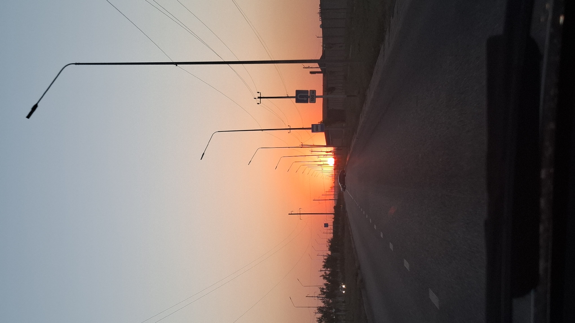 The image depicts a road leading towards a vibrant sunset in Shymkent, Kazakhstan. The sky transitions from a pale blue at the top to a fiery orange and red near the horizon, indicating either sunrise or sunset. A road with white dashed lines marking the lanes stretches into the distance, flanked by numerous streetlights and utility poles with overhead wires. A few vehicles are visible on the road, including one in the foreground and another further ahead. The surrounding landscape appears to be arid, with some sparse vegetation or trees on the left side and a fence or wall on the right. The perspective suggests the photo was taken from inside a vehicle, looking out through the windshield.