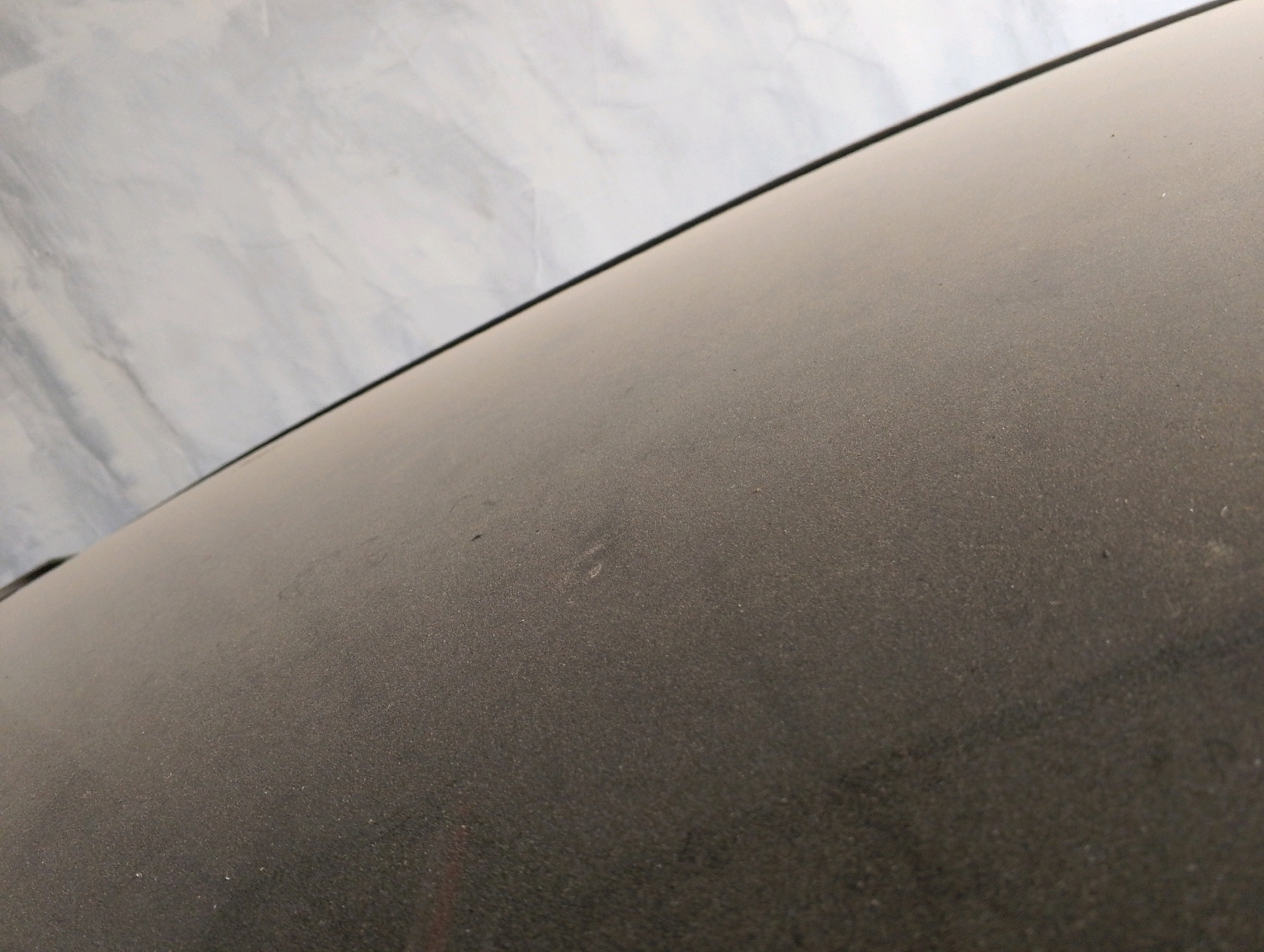 An angled, close-up shot shows a dusty, dark surface, possibly the hood of a car. The surface is covered in a fine layer of dust, with a few very small specks visible. In the background, a wall with a marble-like pattern is visible, with a thin black line dividing the dusty surface from the wall. The scene is devoid of people or any signs of activity. The lighting suggests an indoor setting or an overcast day.