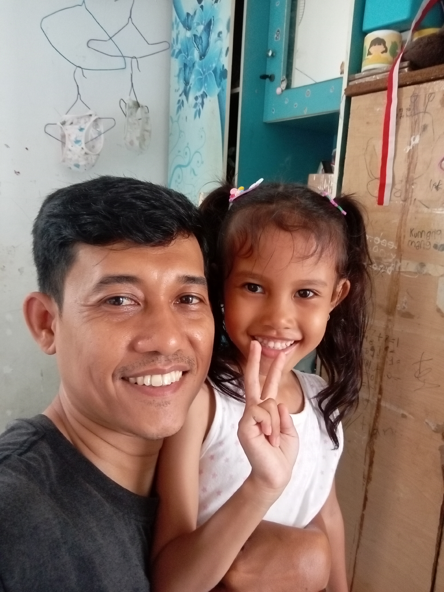 A selfie is captured from a chest-up perspective, featuring a man and a young girl. The man, on the left, has dark hair, a mustache, and a wide smile. He is wearing a dark gray t-shirt. The girl, on the right, has dark hair styled in pigtails with colorful clips and a bright smile. She is wearing a white t-shirt and is holding up two fingers in a peace sign. The background is cluttered, with a wall on the left featuring wire hangers with clothing items. To the right, a wooden surface is visible with some scribbled drawings and text. A bright blue cabinet is also partially visible on the right side. The lighting appears to be natural and somewhat diffused. The image is likely taken indoors in a residential setting in Batam, Indonesia, given the context provided.