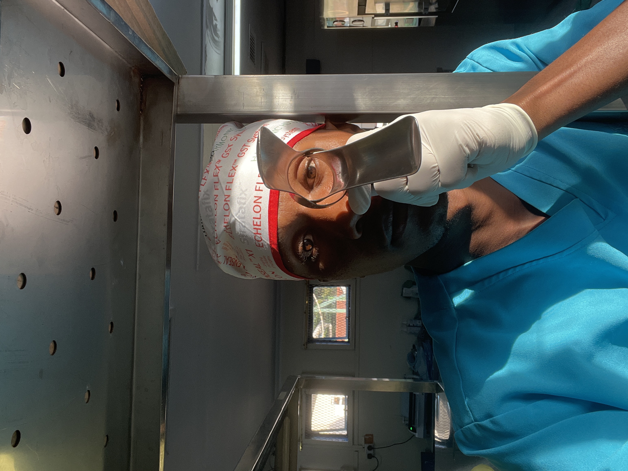 An eye surgery procedure is taking place in Nairobi, Kenya. The surgeon, wearing a blue surgical gown, white cap with red trim, and white gloves, is using a metal surgical instrument to hold open the patient's eye. A metallic stand is located to the left of the patient. The background includes two windows. One has an exterior view of a red brick structure and green vegetation. The other has a view of a white exterior. A surgical instrument cart is visible below the window.