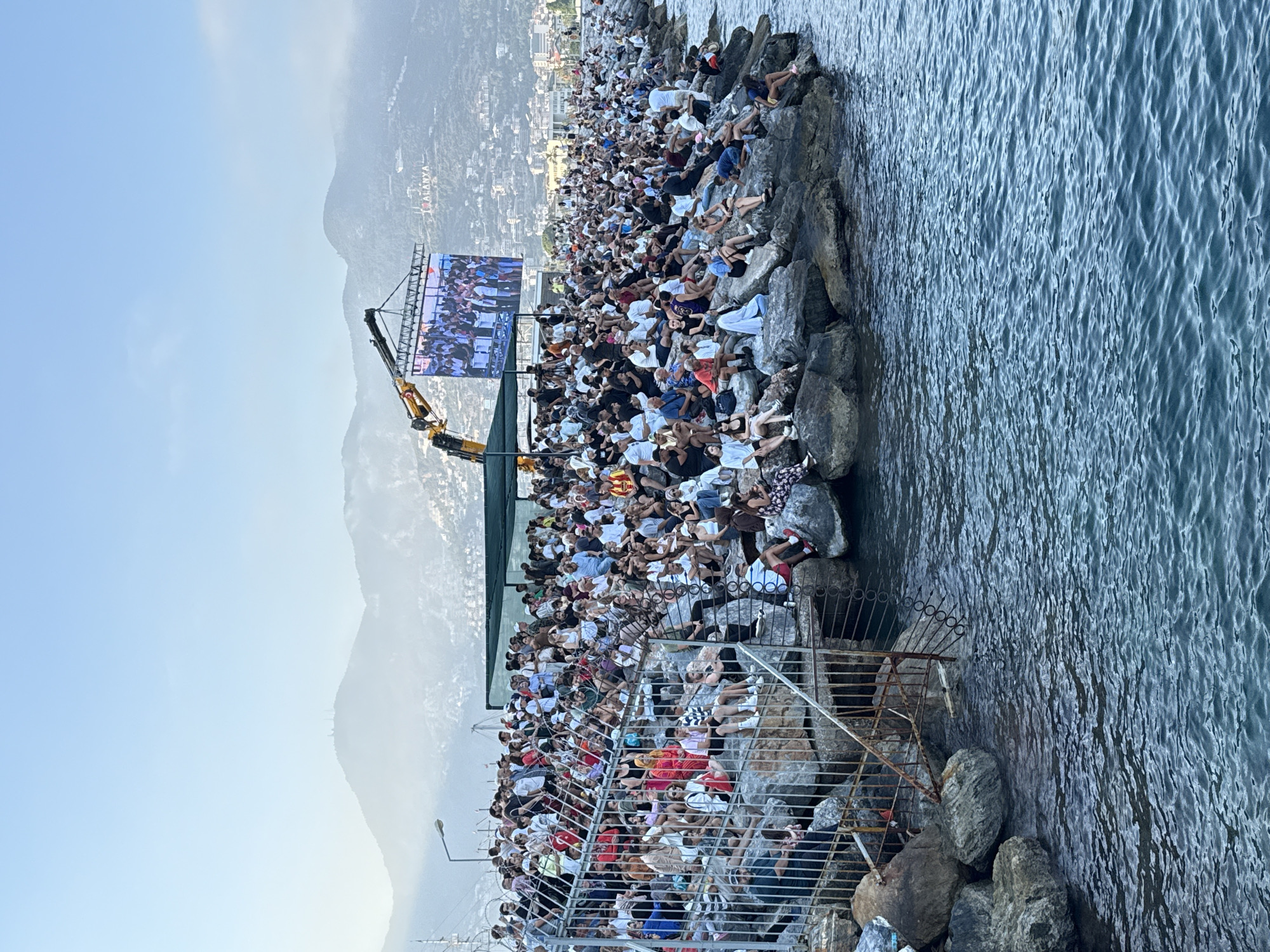 This media file shows a large crowd of people gathered on a rocky coastline, possibly for an event. The scene appears to be set outdoors, with the sea on the right side of the frame and mountains in the background. The sky is clear and light blue, suggesting it is daytime, likely late afternoon or early evening given the soft lighting.
The people are seated and standing on the rocks, facing a large outdoor screen. They are dressed in casual summer clothing, and many appear to be relaxed and engaged with the screen. The crowd is dense, indicating a popular event or gathering. A crane is positioned behind the screen, possibly used for its setup or for other event-related purposes. A metal railing system is visible in the foreground, providing a barrier between some of the audience and the rocky terrain.
The location is identified as Alanya, Turkey, which is suggested by visible text on the screen in the background. The text "ALANYA" is clearly legible, confirming the city. The presence of the sea, mountains, and the rocky coastline are consistent with a coastal location. The overall mood seems to be one of community gathering and shared entertainment.