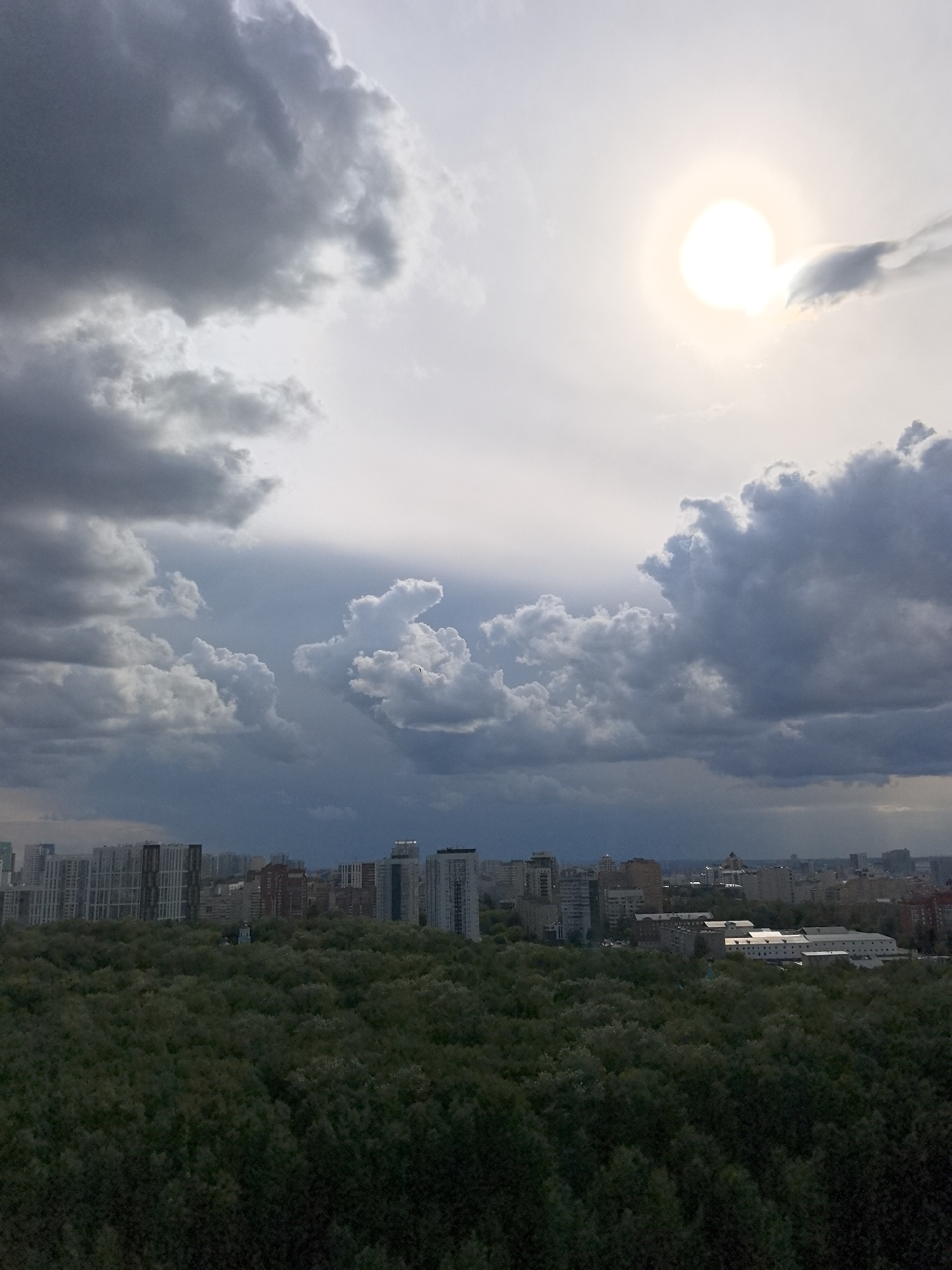 The image depicts a city skyline in Perm, Russia, viewed from a high vantage point, likely an apartment building or rooftop. In the foreground, a dense forest of dark green trees dominates the lower half of the frame. Above the trees, a cityscape with numerous buildings, ranging from residential high-rises to mid-rise structures, stretches across the horizon. The buildings exhibit a variety of architectural styles and colors, with some appearing modern and others older.
The sky above the city is dramatic, filled with large, dark gray storm clouds that cast a somber mood. In the upper right quadrant of the image, a bright, hazy sun shines through a break in the clouds, creating a halo effect. The contrast between the stormy sky and the sun suggests either the beginning or end of a rain shower. No people or specific actions are discernible in the image, making it a static, environmental shot of an urban landscape under a turbulent sky. The overall impression is one of urban development encroaching on a natural environment, under a sky threatening or clearing after a storm.