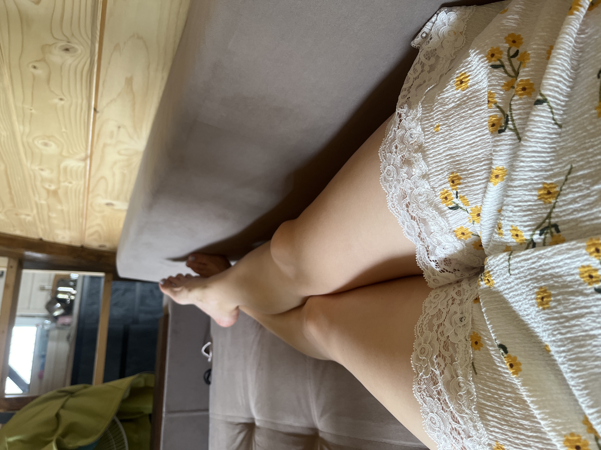 A first-person perspective shot shows a person's legs crossed while resting on a tufted sofa, within a room featuring wooden paneled walls. The person is wearing a white textured dress adorned with a yellow floral pattern and white lace trim. The sofa is a neutral beige color. In the background, a reflective surface, likely a mirror or window, reveals a glimpse of a kitchen area, with cabinets and what appears to be kitchenware. To the left of the reflective surface, a green garment is draped. The overall setting suggests a cozy, indoor environment, possibly a cabin or a home in Mykulychyn, Ukraine, given the prominent wooden architecture.