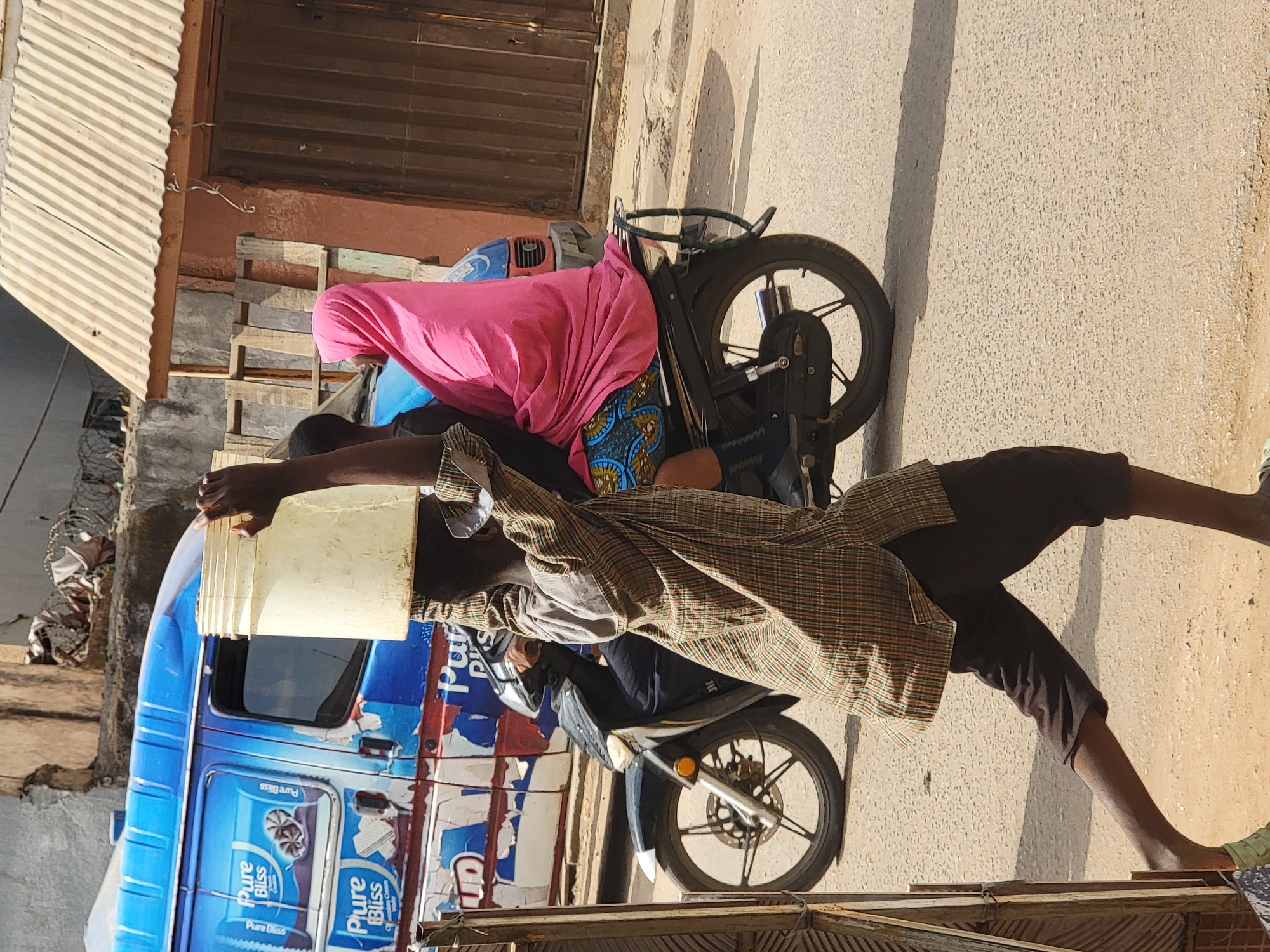 A man is shown refueling his motorcycle on a street corner in Maiburiji, Nigeria. The man is wearing a plaid shirt and dark pants. He is holding a white bucket with a lid over the gas tank of the motorcycle. He is standing next to a blue delivery van with ice cream advertisements. A woman is sitting on the back of the motorcycle. The background shows buildings and a street corner in an urban environment.