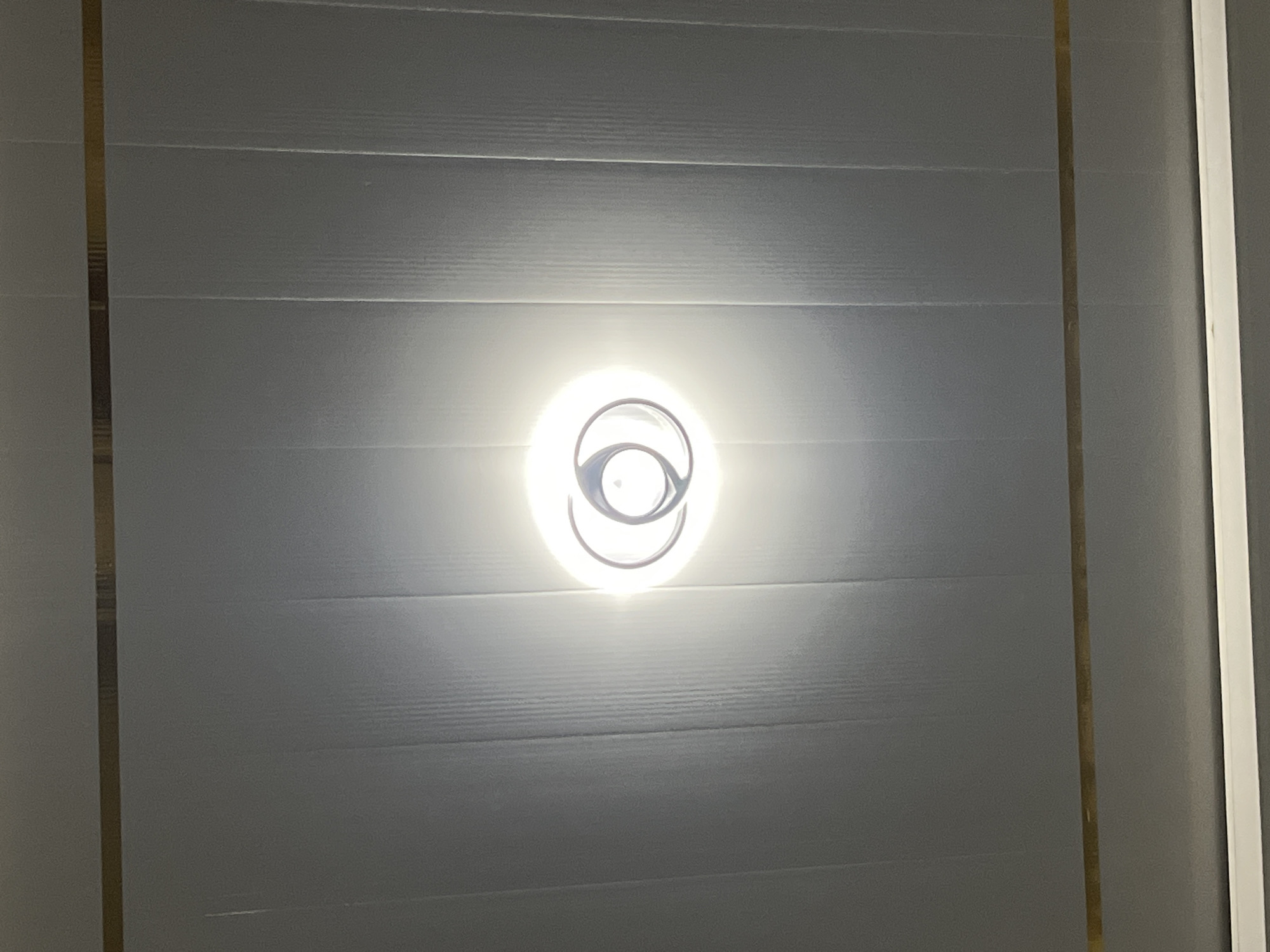 A ceiling light fixture with a circular halo design is illuminated in the center of a white, paneled ceiling. The ceiling has horizontal gold trim bands near the top and bottom. The image appears to be taken from below the ceiling, looking upwards. There are no people or other objects visible in the frame. The location is unknown, and it is not possible to determine if this is in Kebumen, Indonesia.