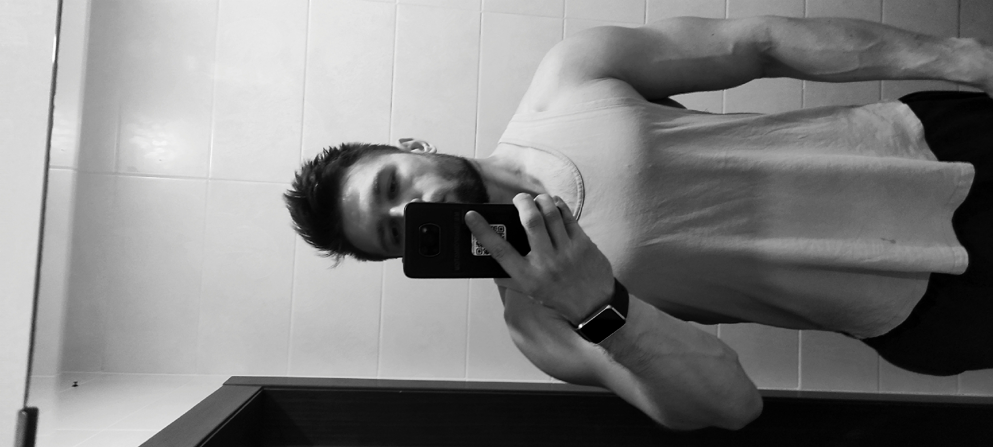 A person stands in a bathroom, holding a black smartphone in their left hand, taking a selfie in a mirror. The person is wearing a light-colored t-shirt and dark shorts or pants. A smartwatch is visible on their left wrist. The bathroom has tiled walls and a dark-colored sink or counter below the mirror. The image is in black and white and is rotated. There's no information to confirm location is in Fryazino, Russia.