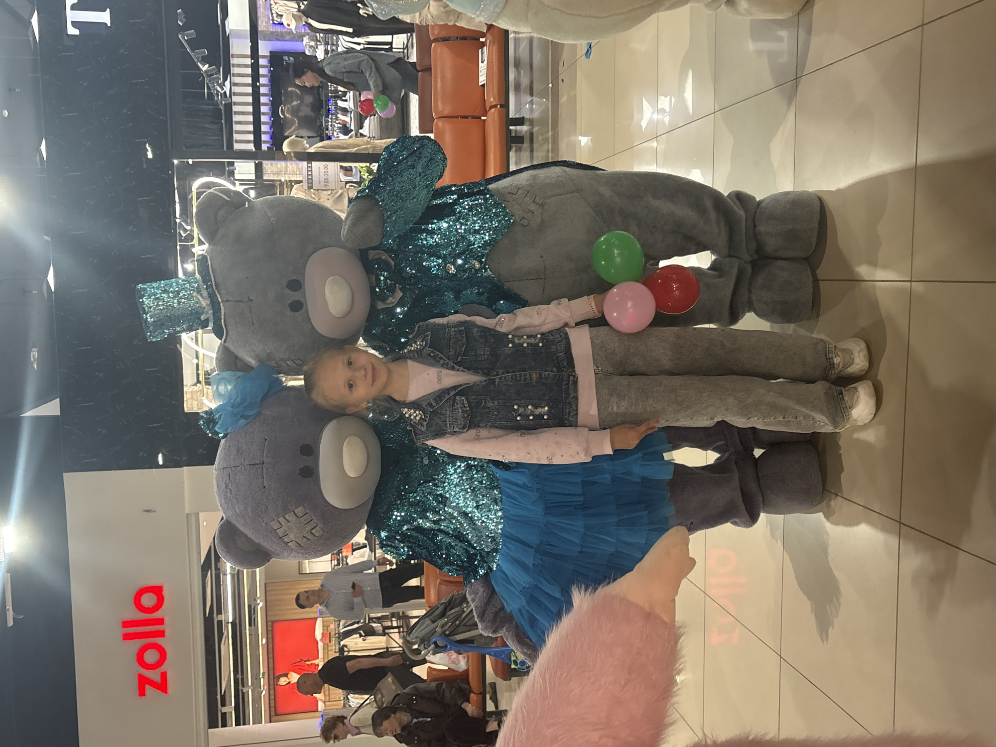 The image shows a young girl posing with two costumed characters resembling bears in a shopping mall or retail store. The bears are dressed in sparkling blue and turquoise jackets. The girl is wearing a denim jacket and grey pants. She is holding three balloons: green, red and pink. In the background is a clothing store, "Zolla," and several other shoppers can be seen browsing. The location is likely within the city of Tri Protoka, Russia, based on the store signage.