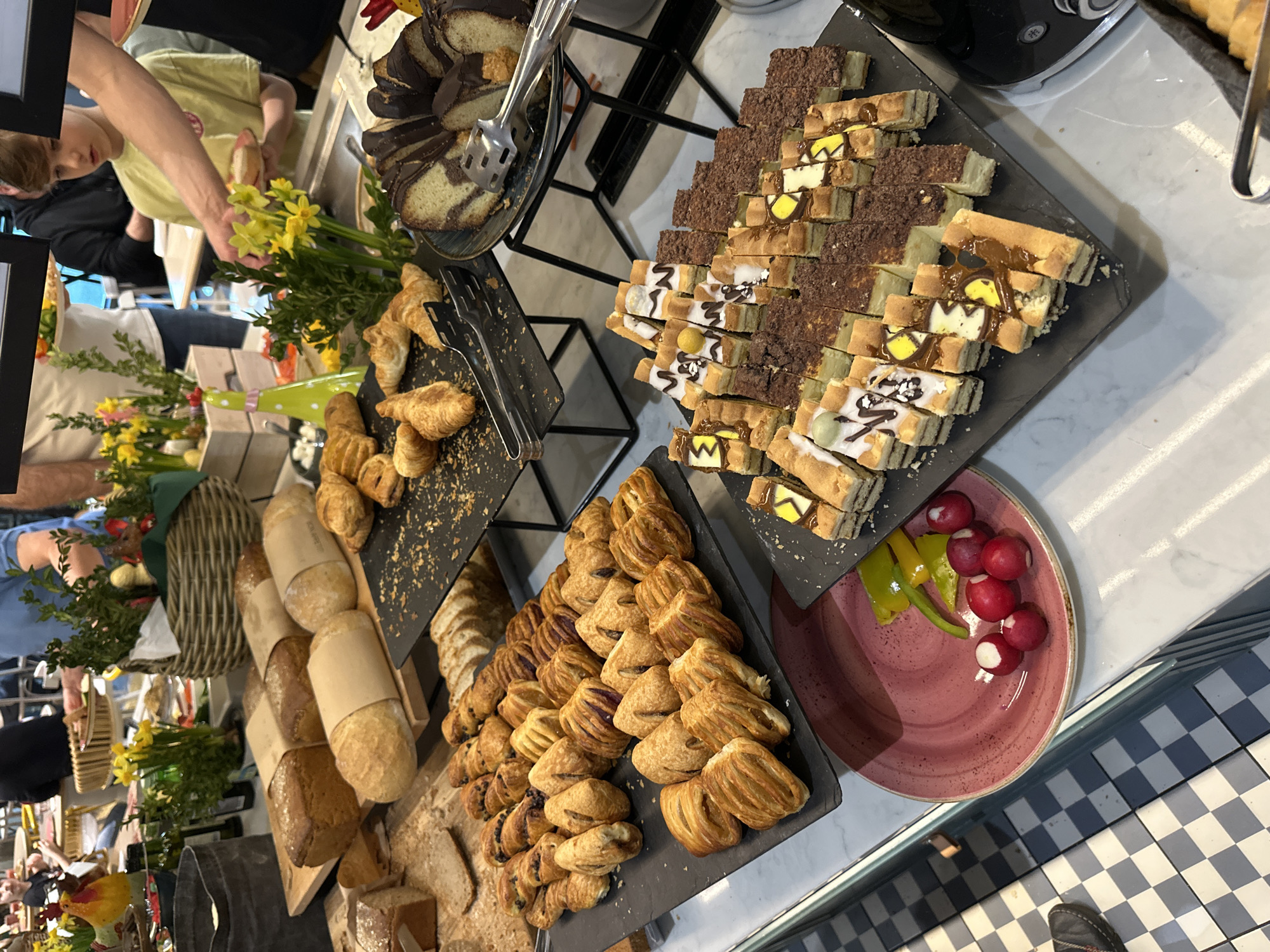 The image captures a buffet-style food presentation, likely in a hotel or restaurant in Wrocław, Poland. The scene features various pastries, breads, and cakes arranged on tiered displays. There are croissants, loaves of bread, chocolate-covered cakes, and intricately decorated pastries. Fresh produce, possibly radishes and peppers, are also present. People are seen selecting food items, and the general atmosphere suggests a breakfast or brunch setting. The environment includes a checkered floor pattern and displays a range of light sources.