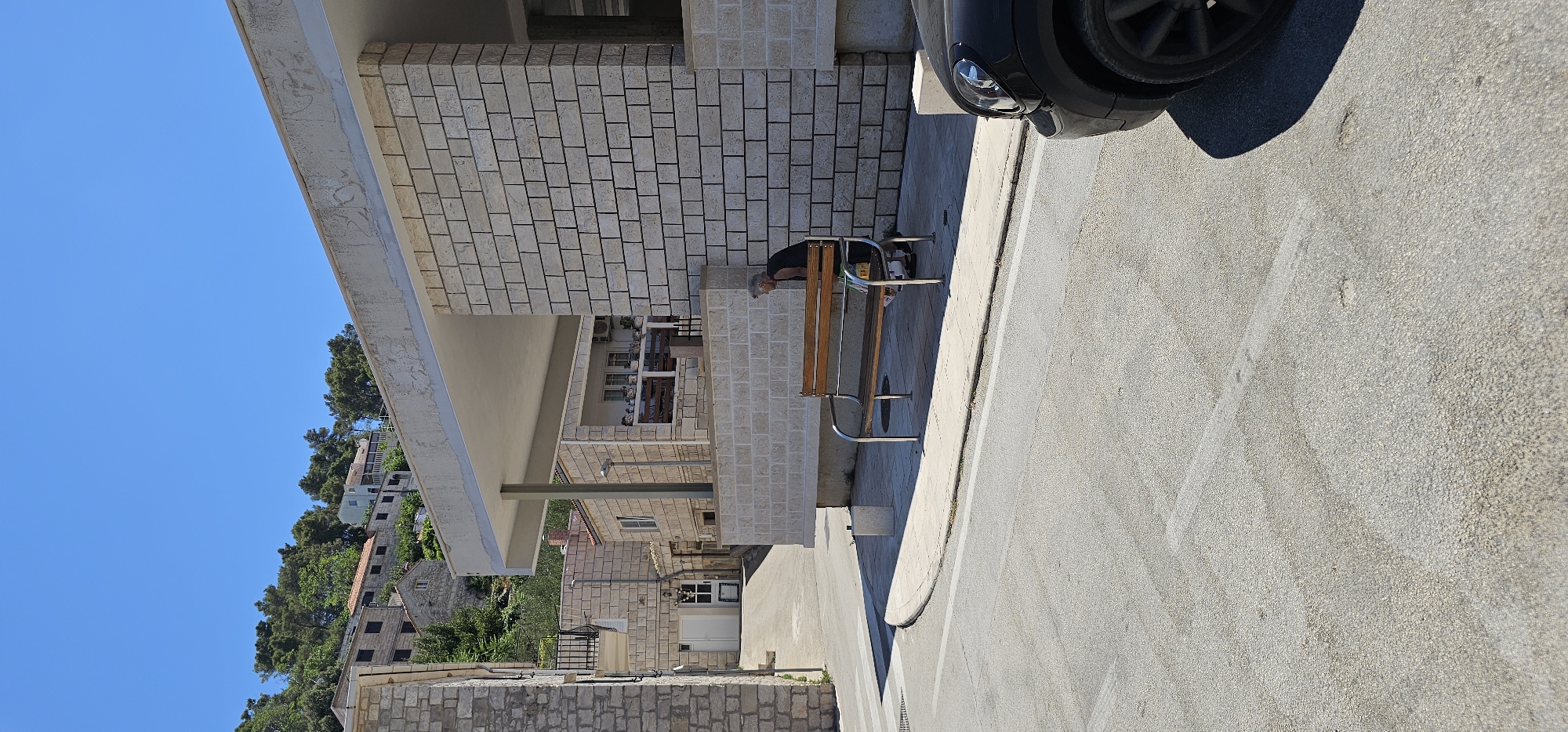 An outdoor, daytime scene captures a person on a bench in front of a stone building. The person, appearing elderly with gray hair, is wearing a black t-shirt and is leaning forward, looking down at something on the bench. The bench itself is made of wood with metal supports. To the right of the bench, a portion of a black car is visible, parked on the street. The environment includes a paved road with white lines, a sidewalk, and a building constructed from light-colored bricks. In the background, more stone buildings and lush green trees are visible on a hillside under a clear blue sky. The location is in Općina Pučišća, Croatia.