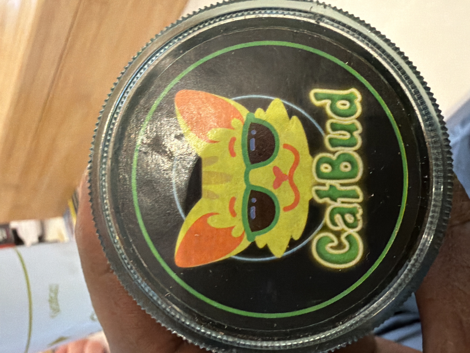 The image presents a close-up, slightly angled view of a circular container lid, held by a person's hand. The lid features a prominent black label with a vibrant central graphic. This graphic depicts a stylized cartoon cat's head, primarily yellow-green with contrasting orange ears and facial features. The cat is wearing bright green-rimmed sunglasses with dark lenses. A bold, bright green circular line borders the central design. Vertically aligned to the right of the cat's head, the text "CatBud" is visible in a playful, bubbly green font with a yellow outline.
The container lid has a metallic-looking, ridged edge, typical of a screw-top design. A person's hand, with light brown skin, is partially visible at the bottom and right, holding the container. The background is blurred, showing a warm, light brown surface, possibly wood, on the left, and indistinct lighter-colored objects with a faint green line on the right. There are no discernible activities beyond holding the container for display. The time of day, weather, and specific location cues (other than the implied indoor setting) cannot be determined from the image itself, though the context provided is London, United Kingdom. No specific emotions are conveyed.