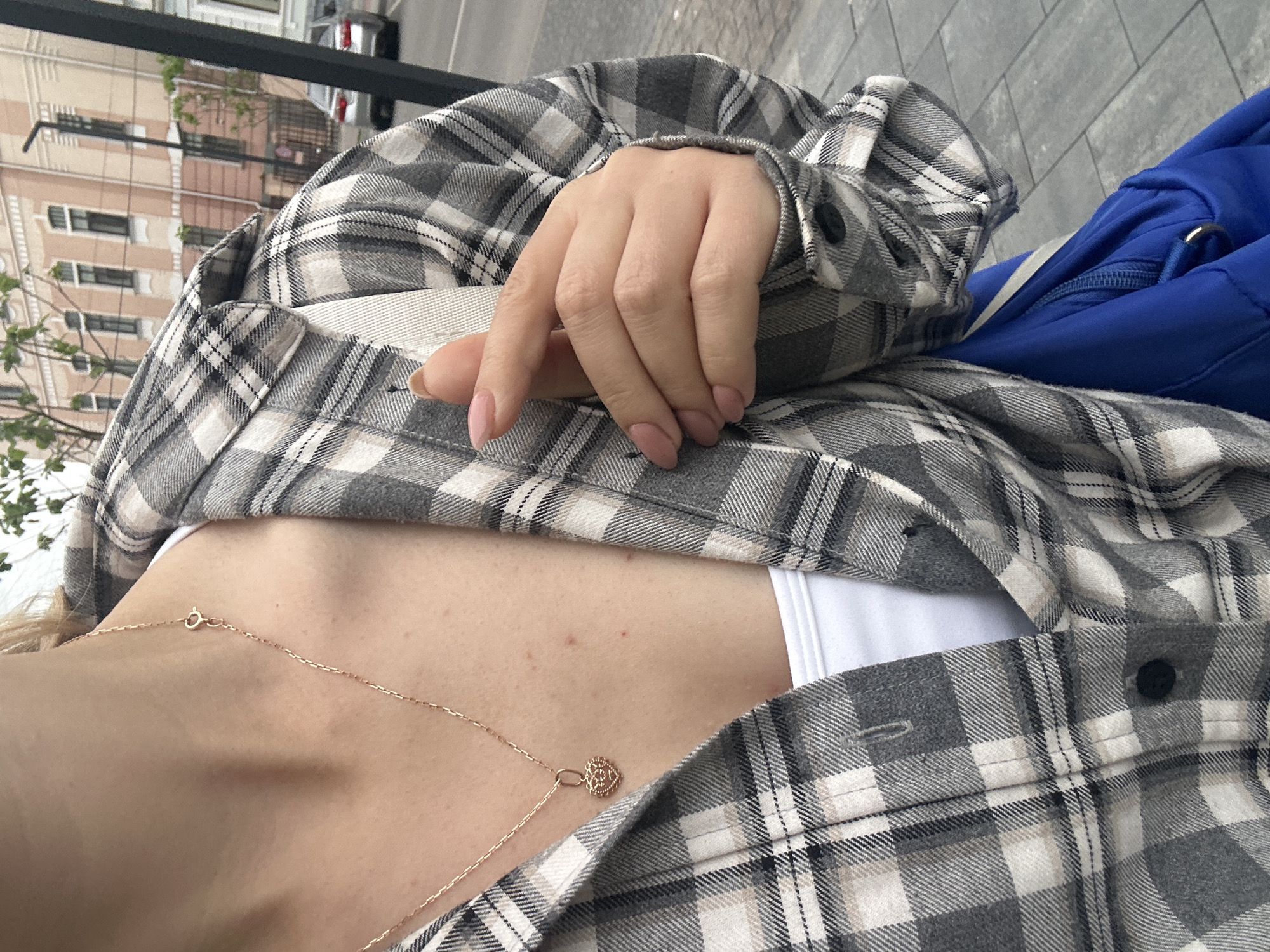 A close-up image captures a person, likely female, from the chest to just above the waist, situated outdoors on an urban street in Ternopil, Ukraine. The subject is wearing an unbuttoned grey, white, and tan plaid flannel shirt over a white tank top or camisole. A thin, rose gold-toned chain necklace with a small, ornate heart-shaped pendant hangs around the neck; its spring-ring clasp is visible near the upper right clavicle. Several small, red blemishes are present on the visible skin of the chest. The person's right hand, featuring light pink manicured, almond-shaped fingernails, rests casually on the plaid shirt in the mid-right portion of the frame. A portion of a bright blue fabric bag, possibly a backpack, with a silver zipper pull is visible in the lower right corner, partially obscured by the shirt. The background reveals a city street with grey paving stones, a large multi-story building with a light peach-colored facade and dark windows, green trees, and two silver sedans parked in the distance. A black metal pole or railing is visible on the far right edge of the image.