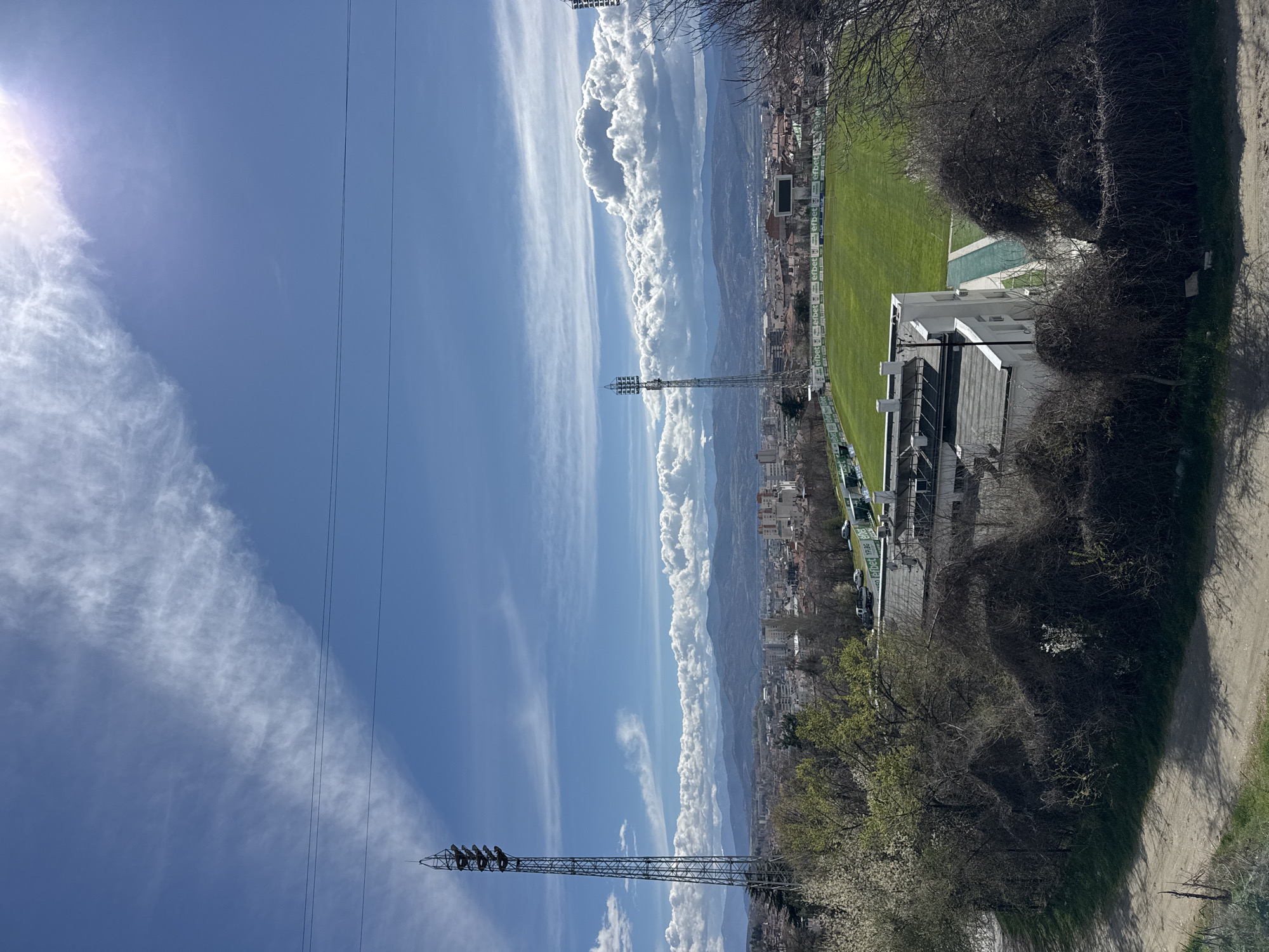 The image depicts a view of the city of Blagoevgrad, Bulgaria, and a stadium complex on a sunny day. A football stadium with a green field and adjacent buildings and pool is visible. The stadium is surrounded by urban development, and mountains can be seen in the distance. Two metal utility towers stand in the foreground. Power lines stretch across the sky. The sky is blue with elongated clouds. There are trees along the lower right side of the photo.
