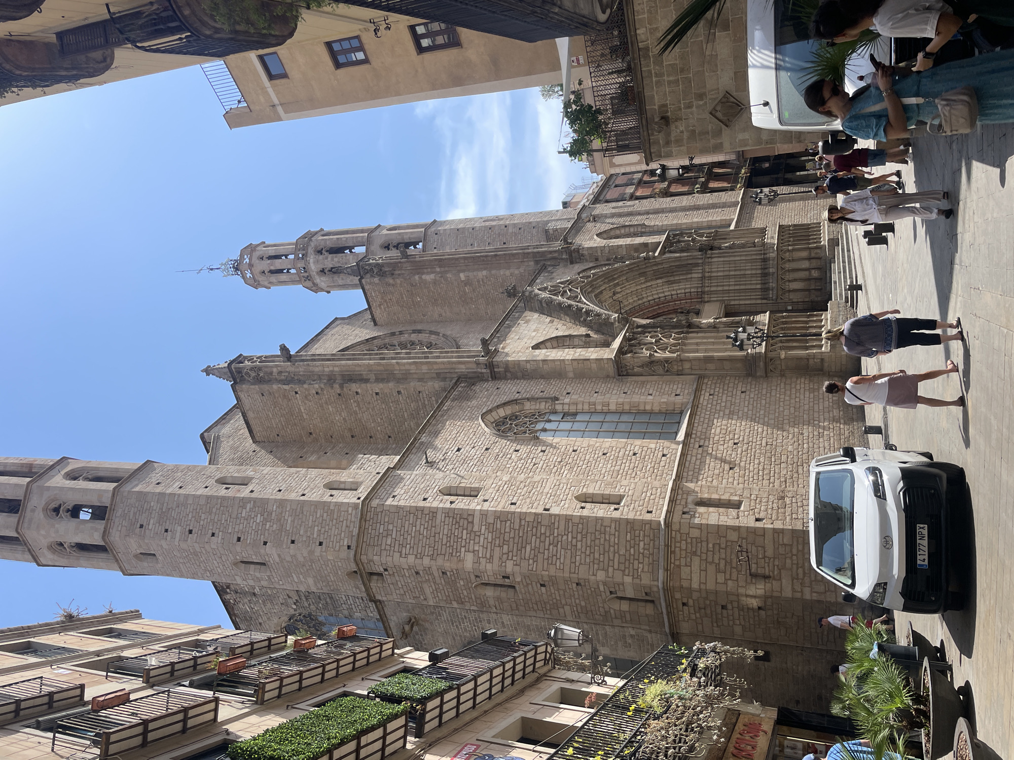 The image depicts a sunlit, narrow street in Barcelona, Spain, leading towards a large, Gothic-style stone church. The church features a prominent bell tower and an ornate arched entrance. To the left of the church, a multi-story building with balconies and lush green plants is visible. On the right side of the street, another building of similar height with windows and balconies is present. A white van is parked in the street, and several people are walking in the foreground and middle ground, some looking at their phones. The sky is clear blue, indicating a sunny day.