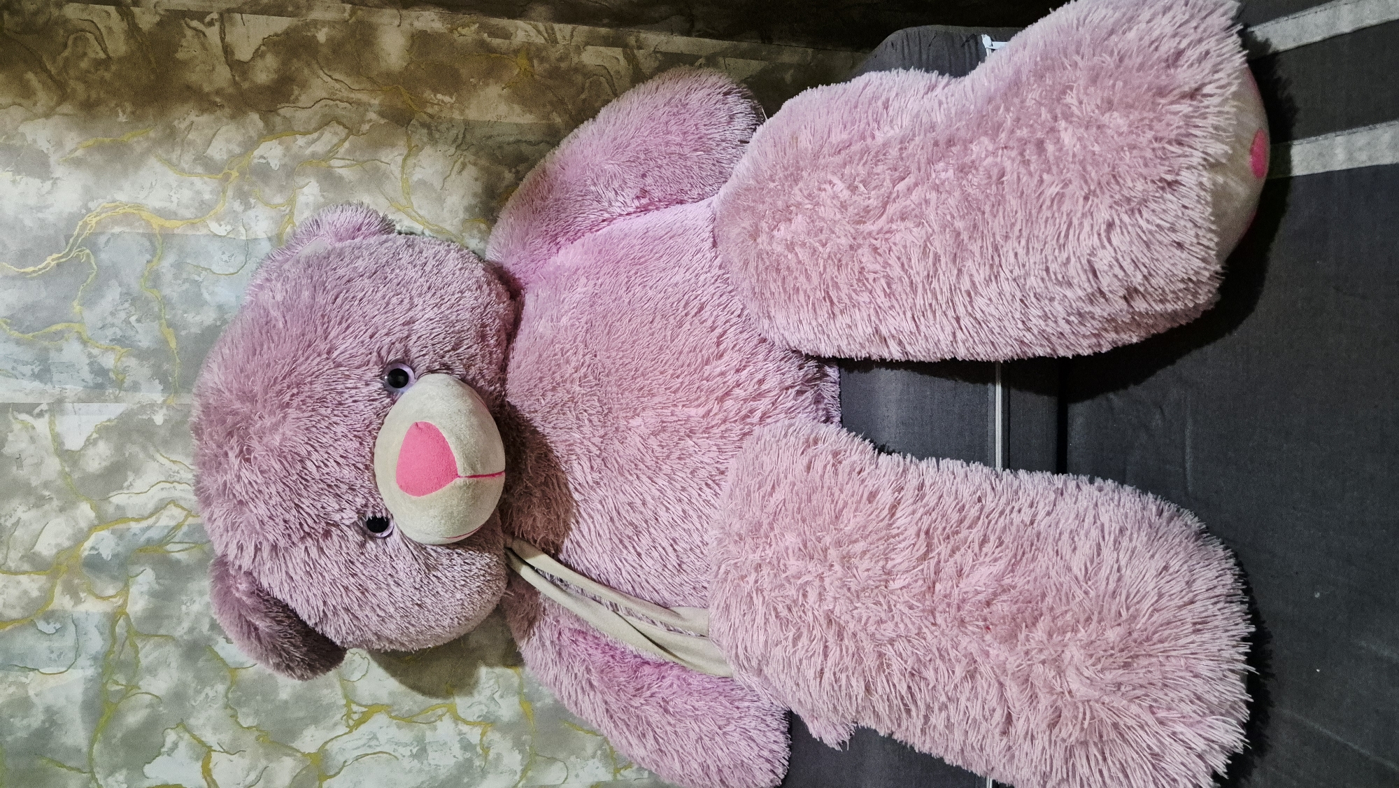 A large, plush, lavender-colored teddy bear is seated on a dark gray sofa. The bear has a light beige snout with a pink heart-shaped nose and dark eyes. A beige ribbon is draped around its neck. The background features a textured wallpaper with a marbled pattern of gray, white, and gold lines. The lighting is even, with some shadows cast by the bear on the sofa and the wall. The scene is indoors and appears to be a residential setting. There is no visible indication of the city of Karawang, Indonesia.