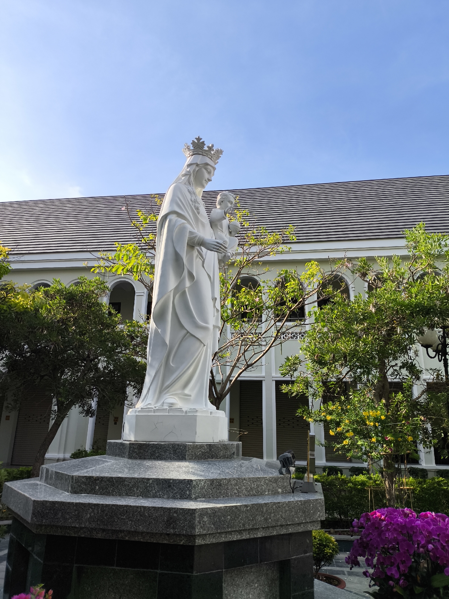 The image shows a white marble statue of the Virgin Mary holding the infant Jesus, set on a multi-tiered granite pedestal. The statue is positioned outdoors in front of a building with a grey tiled roof and white walls featuring arched windows. Lush green trees and bushes surround the statue, with a cluster of vibrant purple orchids in the foreground. The sky above is clear blue. The scene appears to be in a tranquil setting, possibly a church courtyard or garden, located in Bac Nha Trang Ward, Vietnam.