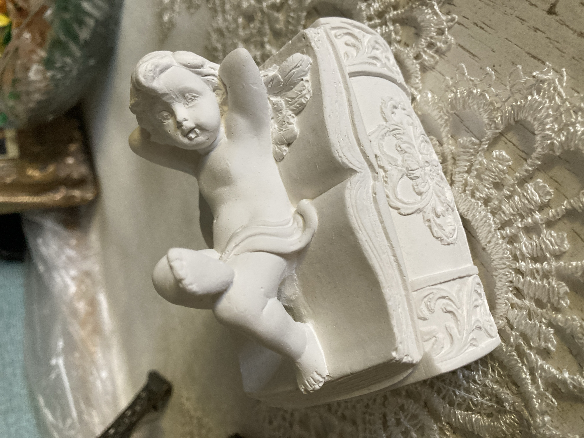 A white, plaster figurine of a cherub is lying on its side atop an open book, which serves as the lid of a box. The cherub is depicted with its head tilted back and to the right, its mouth slightly open, and its eyes looking upwards. One leg is bent, and the other is extended. The book is intricately detailed with embossed scrollwork on the sides. The figurine and box are placed on a lace-covered surface, which is set against a blurred background of other objects. The scene appears to be indoors, in an unknown location within an unknown city.