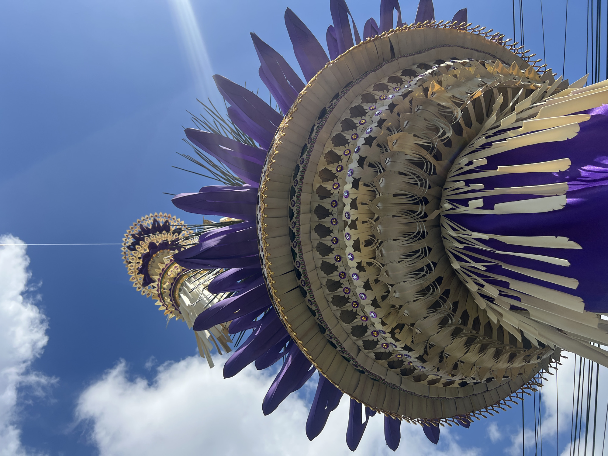 A low-angle, eye-level shot captures a decorative Balinese umbrella, likely made of woven palm leaves and adorned with purple fabric and gold trim, against a bright blue sky with scattered white clouds. The umbrella, positioned in North Kuta, Indonesia, is the central subject, with its intricate layers and decorative elements prominently displayed. Sunlight glints off the upper portions of the umbrella and creates a lens flare in the upper right corner of the frame. The perspective emphasizes the height and grandeur of the decoration, which appears to be suspended, possibly for a religious ceremony or festival. The lower portion of the image shows a glimpse of overhead electrical wires, indicating an urban or semi-urban setting. No people are visible in the frame, and no actions or interactions are depicted. The focus is solely on the ornate umbrella and its stark contrast against the natural backdrop of the sky.
