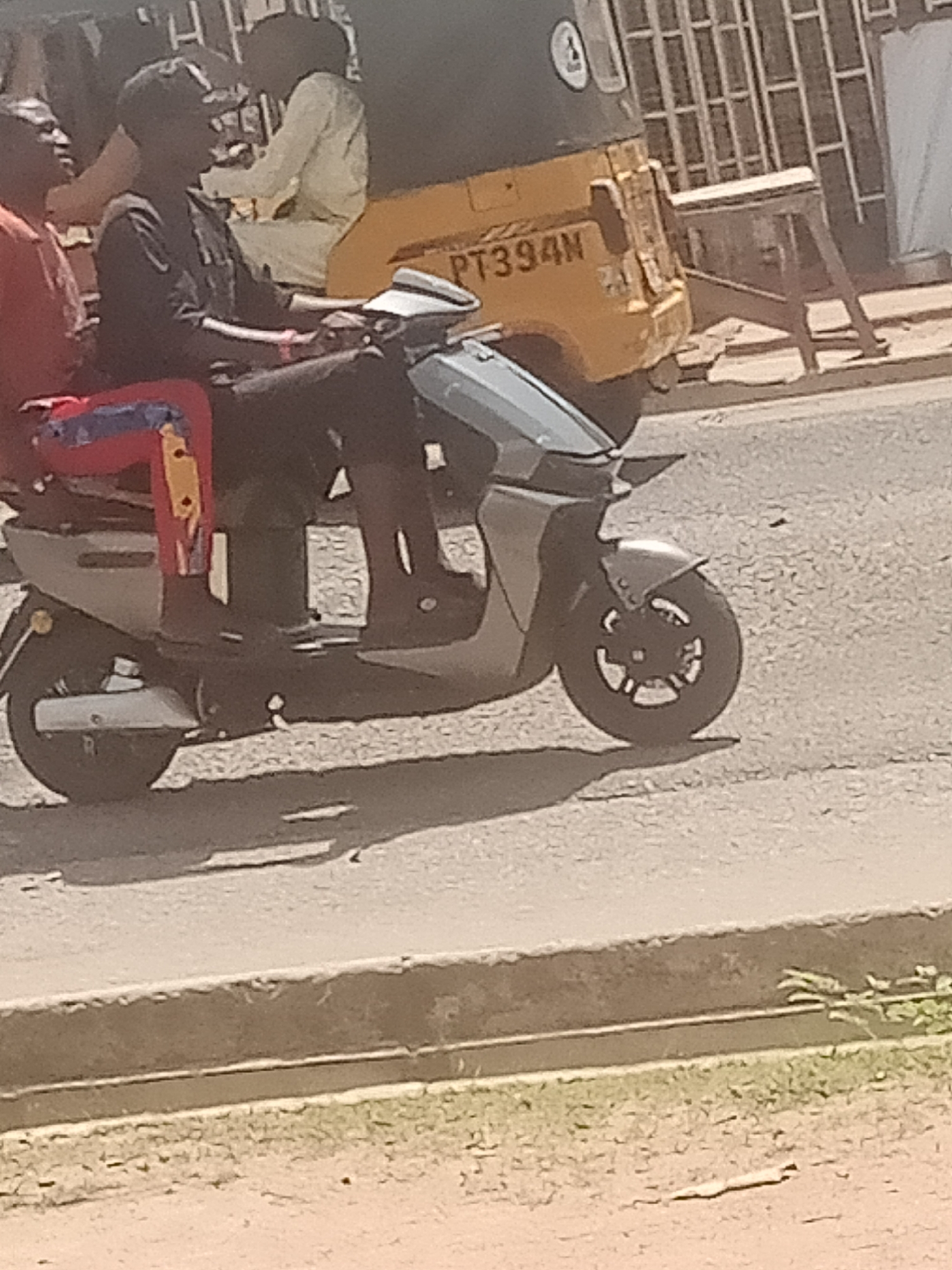 The image depicts a street scene in Jalingo, Nigeria. A man is riding a scooter, with another person seated behind him. The rider is wearing dark clothing and a helmet. The passenger is wearing a red garment with blue and yellow accents, and a yellow object is visible on their lap.
Behind the scooter, a yellow tricycle is partially visible. The tricycle has a license plate with the text "PT394N". The background shows a building with a barred window and what appears to be a wooden structure.
The scene appears to be taking place during the daytime, with sunlight casting shadows on the road. The road surface is paved, and there is a dirt or gravel area in the foreground with some sparse vegetation. The overall impression is of everyday activity on a street in a Nigerian town. No specific emotions are discernible from the subjects.