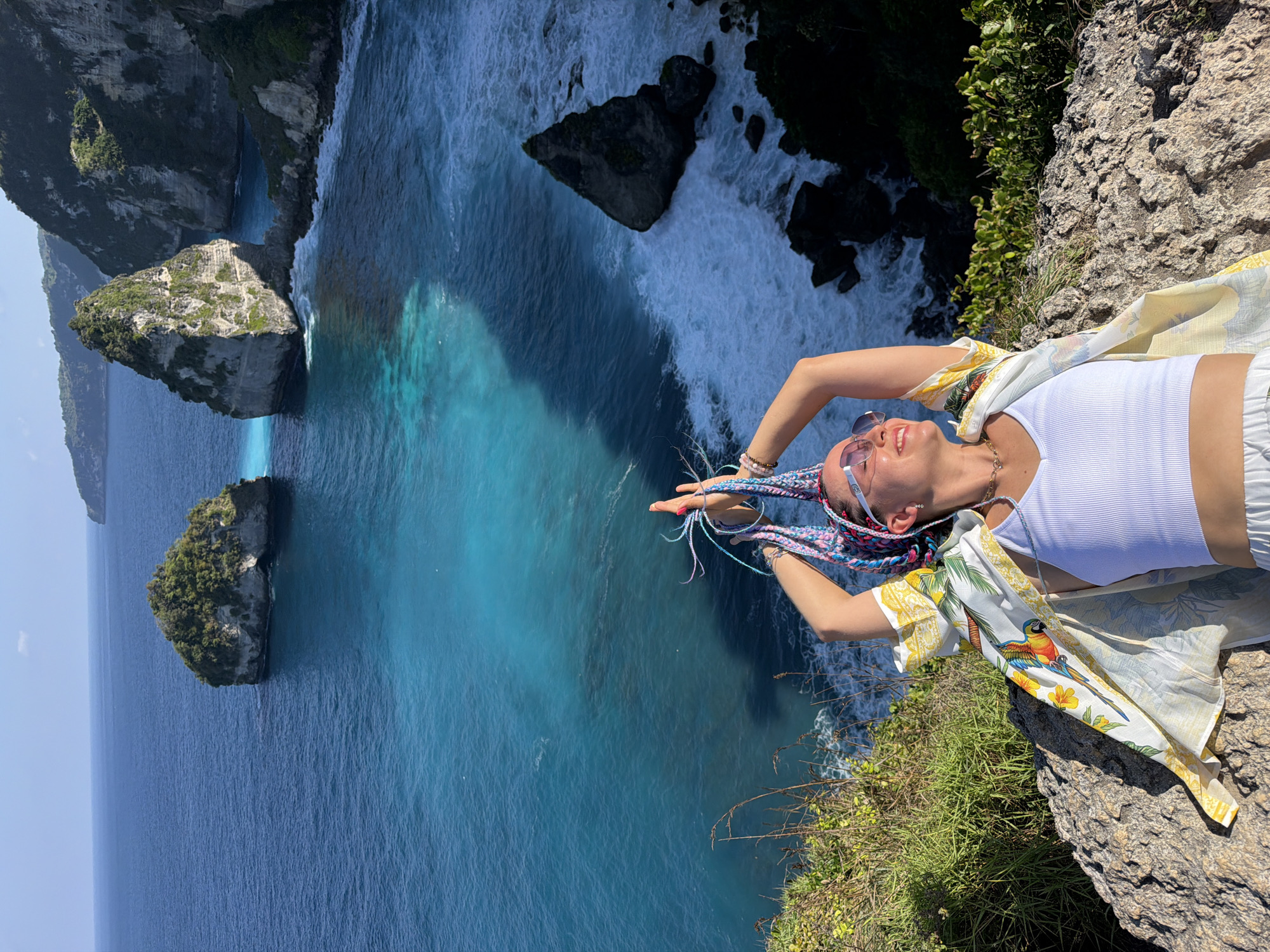 A woman with blonde braided hair is lying on a cliff edge overlooking a turquoise ocean in Nusa Penida, Indonesia. She is wearing a white tank top, white pants, and a white open shirt with a parrot and floral print. She has sunglasses on and her arms are raised above her head. The ocean has several rock formations and waves crashing on the shore. The sky is clear and blue.
