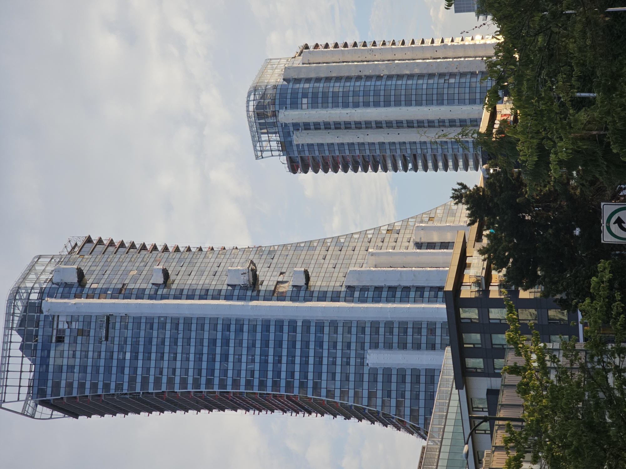 The image shows a cityscape featuring two prominent modern high-rise buildings under construction in Vancouver, Canada. The building on the left is a very tall, sleek skyscraper with a glass facade, featuring scaffolding and construction equipment on its exterior, suggesting ongoing development. It has a unique curved shape and a prominent steel framework at its peak. The building on the right is also a high-rise, with a rounded facade and multiple balconies. It too shows signs of construction, with white coverings on vertical elements and scaffolding on its sides. In the foreground, there are trees and a low-rise building with dark-colored panels and balconies, also appearing to be residential. The sky is overcast with some visible clouds. The overall scene depicts a growing urban environment in Vancouver, characterized by contemporary architecture and ongoing construction projects.