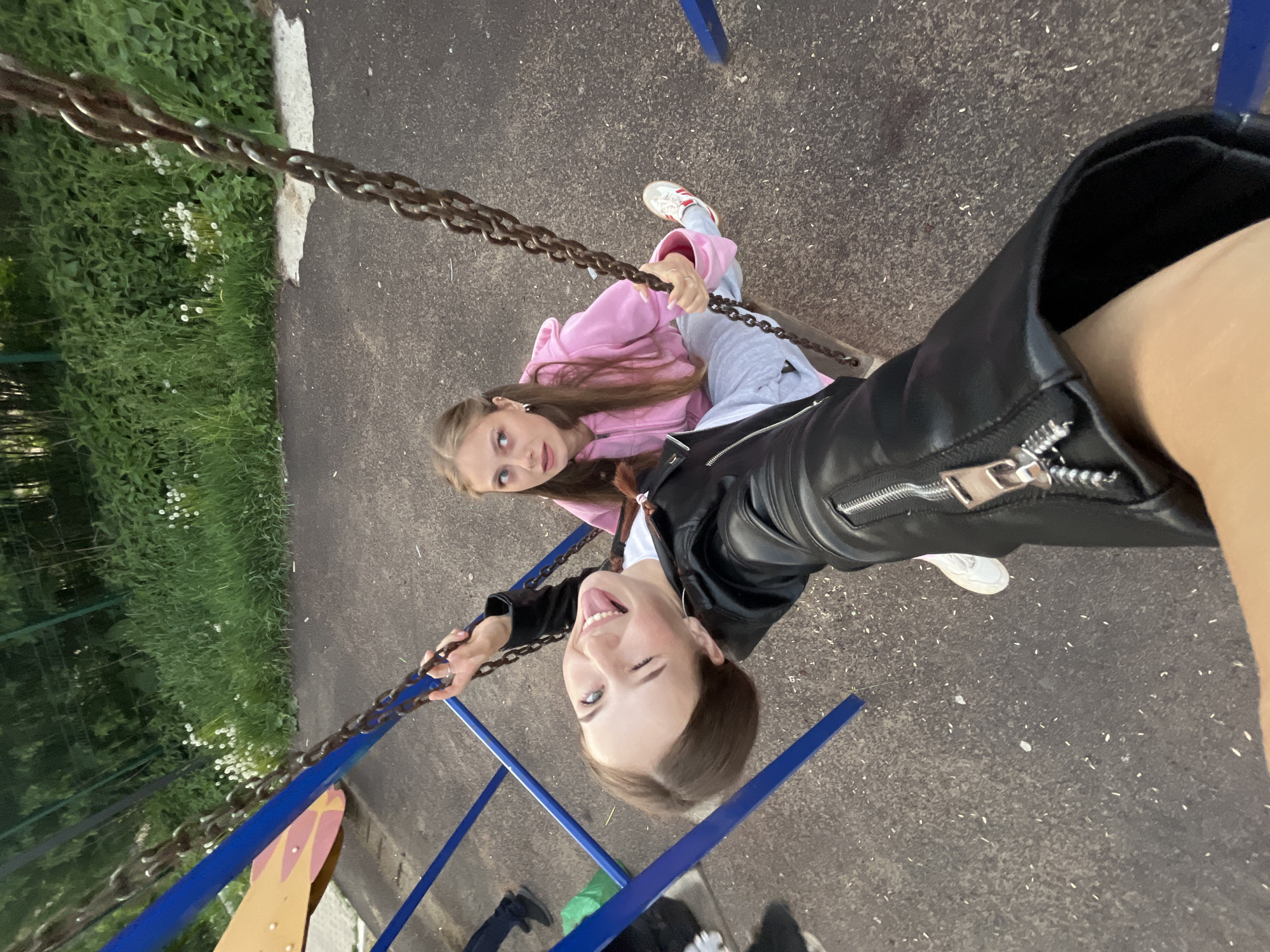 Two young women are on a swing set in what appears to be a public park or playground. The woman closest to the camera is wearing a black leather jacket and sticking her tongue out, winking as she holds the swing chain, implying she is swinging. The other woman is wearing a pink hoodie and grey sweatpants, holding onto the chains with an inquisitive expression. The swings are blue. There is greenery and a sidewalk nearby. The setting suggests an urban area, potentially in the city of Фоки, Russia.