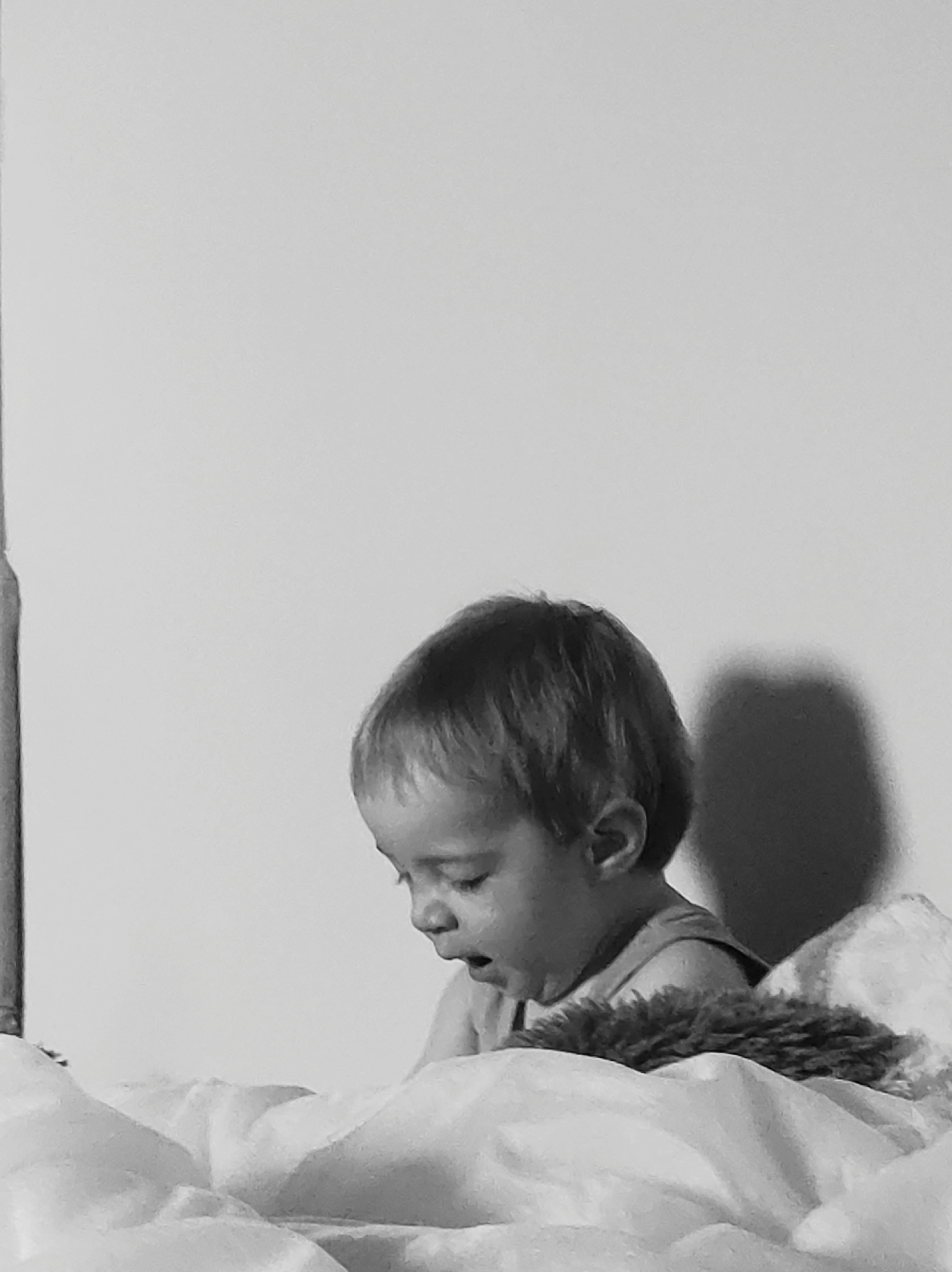 A black and white photograph shows a child sitting on a bed or possibly a couch. The child appears to be looking down, possibly at something in their hands or on the bed. The child is wearing a tank top. There is a fluffy object that may be a toy or a pillow, next to the child. The background is a plain white wall, with a shadow of the child. The location is Unknown, Unknown.