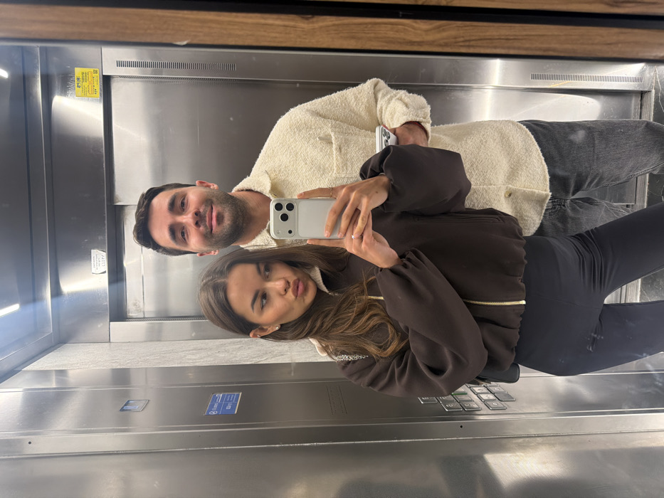 The image depicts two individuals, a female and a male, taking a selfie reflected in a mirror within a modern elevator. The elevator interior features polished stainless steel walls and an illuminated control panel on the left, displaying multiple buttons, a digital readout, and blue informational plaques indicating "Max. 6 Persons / 450 kg" capacity and an emergency phone icon. A yellow sticker with Hebrew text, partially visible on the rear wall's reflection, indicates an 'Error code: 1121' and instructs to 'Please call a technician,' confirming the location in Israel, consistent with a building in Tel-Aviv.
The female, positioned in the foreground and slightly to the left, has long, dark brown hair and is wearing a dark brown zip-up jacket over dark leggings. A silver chain strap, likely from a bag, is visible over her left shoulder. She holds a light-colored smartphone with a distinctive triple-lens camera module in her left hand, on which a ring is visible on her ring finger, and poses with a pouting expression while looking into the reflection. The male, standing behind and to the right of the female, has short dark hair and stubble. He is wearing a cream-colored, textured long-sleeved collared shirt or jacket and dark jeans. He holds a white object in his right hand and displays a faint smile, also looking towards the reflection. The overall lighting is bright and even, likely from internal elevator fixtures.