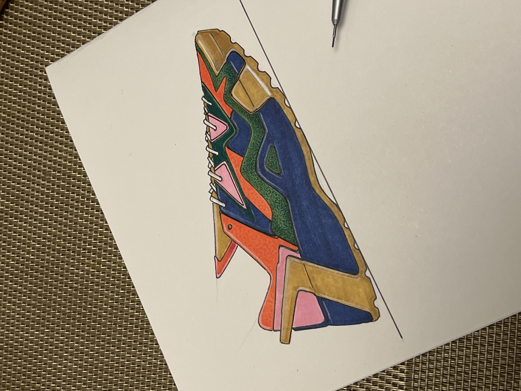 The image depicts a drawing of an abstract object with geometric shapes filled in with different colors: blue, green, orange, pink, and gold. The drawing is on a piece of white paper which is lying on a brown woven surface. A mechanical pencil is positioned at the top right corner of the paper. Two diagonal lines are sketched from the top to the bottom of the paper.