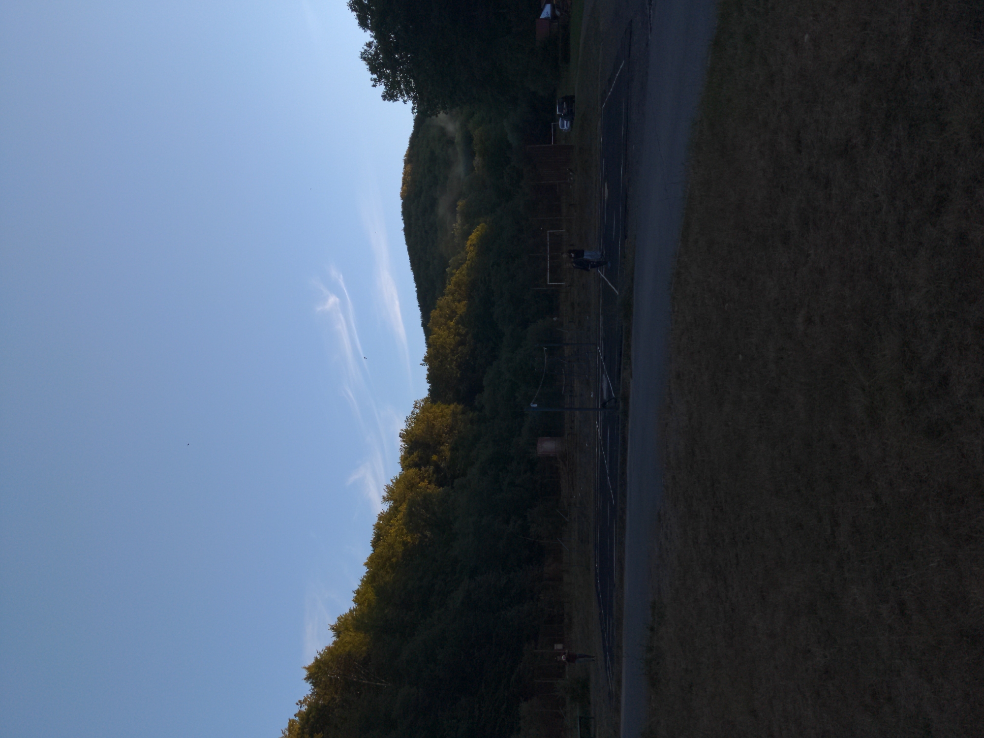 A medium shot captures an outdoor scene with a clear blue sky above a wooded hill. The hill is densely covered in dark green trees, with some areas illuminated by sunlight, creating a contrast between shadow and light. A few wispy clouds are visible in the sky. In the foreground, a dark asphalt surface marks what appears to be a sports court or parking area, with faint lines indicating boundaries. A grassy area with sparse vegetation is in the immediate foreground, mostly in shadow. Two people are standing on the dark surface, one appearing to be walking. Further back, there are some structures, including a net that could be part of a volleyball court, and a fence. In the distance, a few vehicles are parked. The overall impression is of a somewhat secluded outdoor recreational area, possibly part of a park or community space, situated at the base of a hilly, forested landscape.
