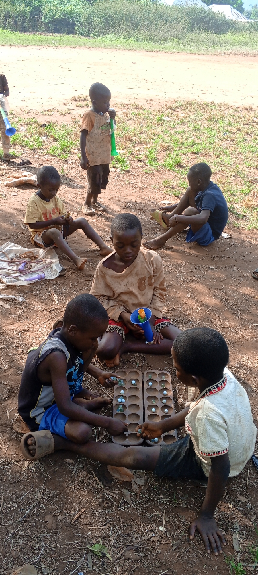 A group of six children are sitting on the ground in Ngalula, Democratic Republic of the Congo. Two children are playing a board game with marbles, while the others look on. One boy has a blue and green toy horn. The ground is dirt with some patches of grass.