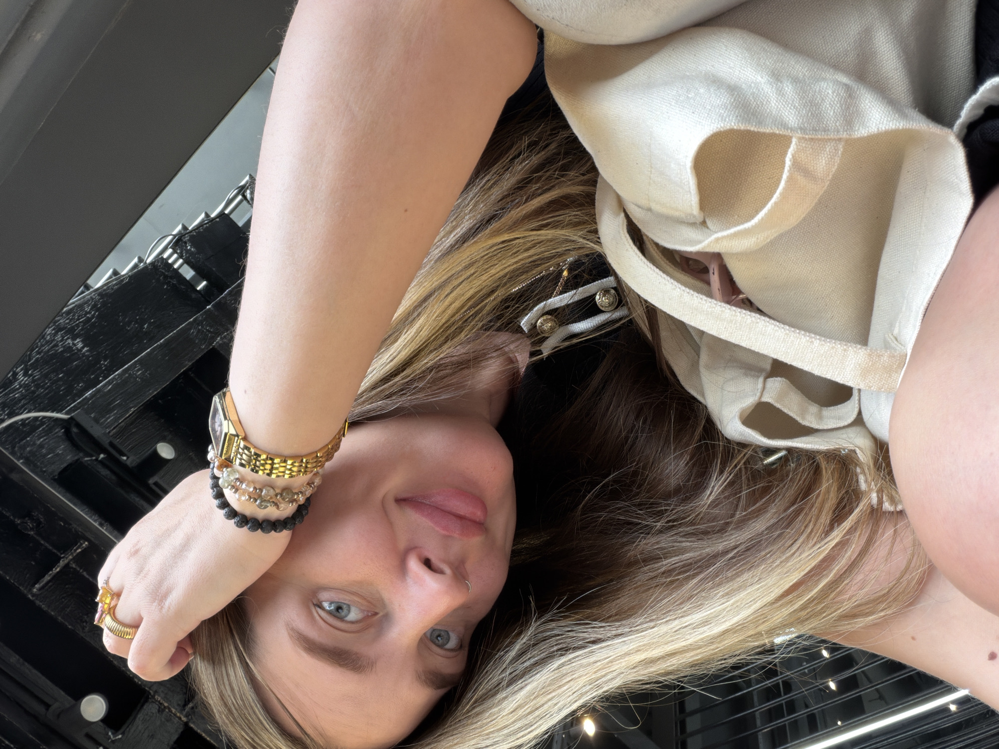 A selfie of a woman with blonde hair, fair skin, and blue eyes wearing a gold watch and multiple bracelets is tilted at an unusual angle. She is wearing a nose ring and has rings on her fingers. A light-colored tote bag is slung over her shoulder. The background appears to be the ceiling of a building with black metal beams and lighting fixtures. Based on the style of the metalwork, it resembles the Moscow metro station Mayakovskaya, located in Moscow, Russia.