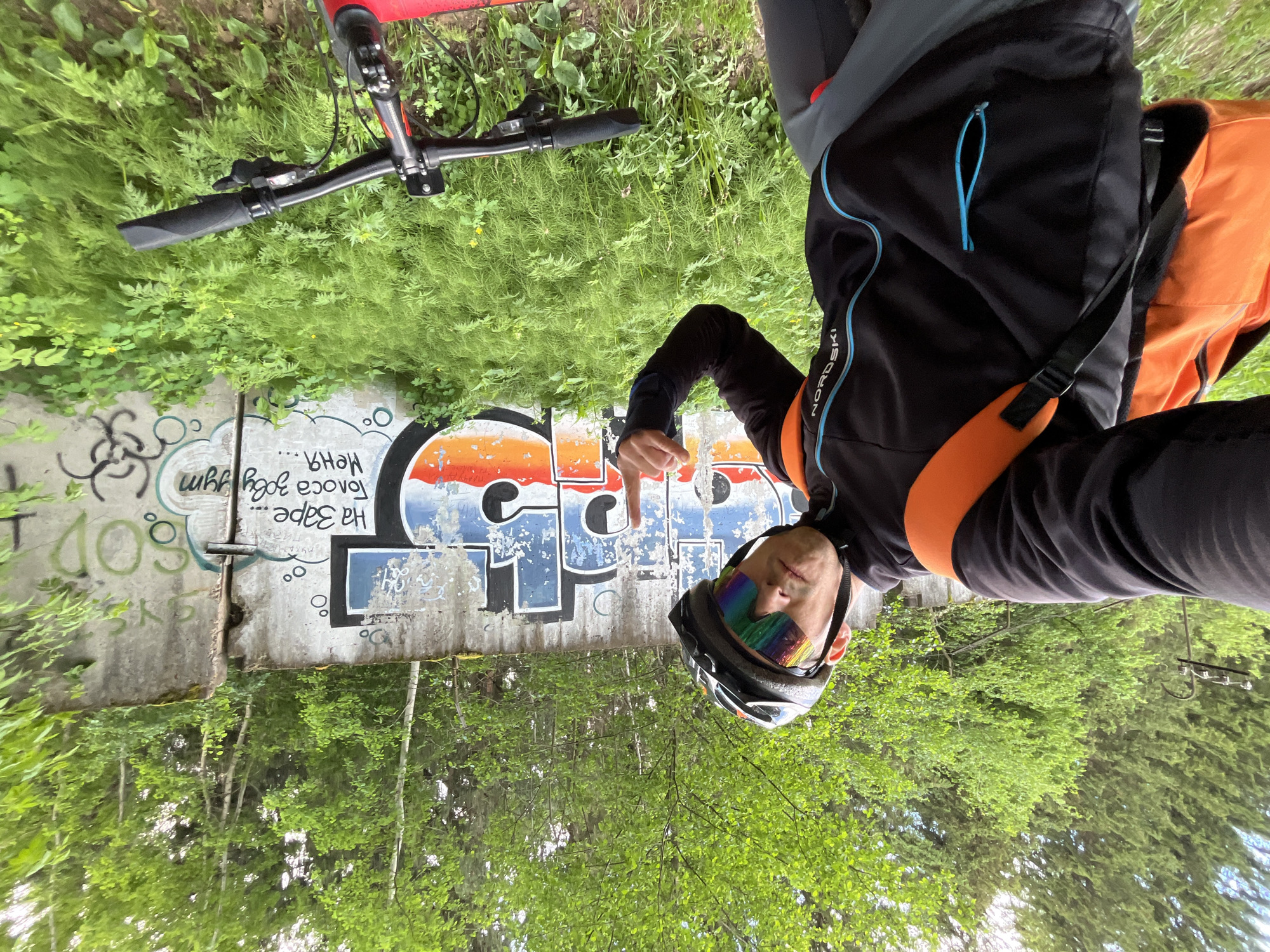 An outdoor, vertically oriented selfie captures a cyclist posing in what appears to be Chaikovsky, Russia. The individual, seen from the chest up, wears a black long-sleeved cycling jersey with light blue accents, a black cycling helmet, and a striking rainbow-colored visor that covers their eyes. An orange chest strap or bag strap is visible across their torso. The cyclist's right arm is bent at the elbow, with the index finger pointing towards the upper right of the frame, giving a casual or assertive pose.
Behind the cyclist is a large concrete wall or barrier, heavily covered in graffiti. Prominently featured on the wall is a large, colorful graffiti piece with block letters, some of which appear to form Cyrillic words. The letters 'P' and 'T' are clearly discernible, possibly part of "СПОРТ" (SPORT). Other smaller inscriptions are visible, including the Cyrillic phrase "На Заре Голоса Звучат" (Voices Sound at Dawn) and the word "меня" (me/my), along with English text "God" and various abstract symbols and circles.
To the upper left of the person, part of a red bicycle's frame and handlebars are visible, partially obscured by green foliage. Lush green grass, bushes, and trees densely fill the foreground and background, suggesting a natural or semi-natural setting. The overall lighting indicates daytime, likely a bright or overcast day. The jersey also displays the brand "NORD SKI" on its side. The scene conveys a moment of outdoor activity and self-expression amidst both urban art and nature.