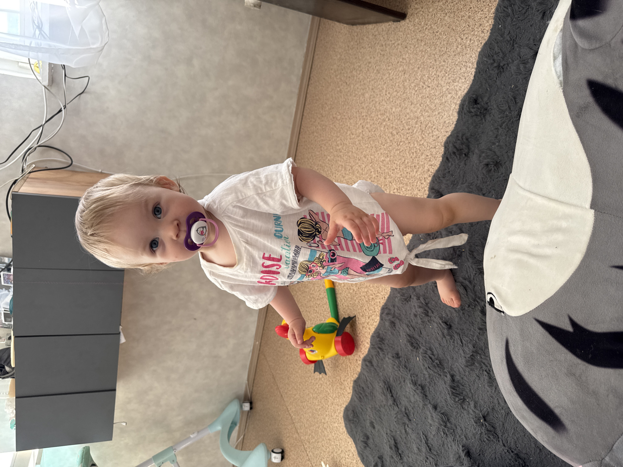 A toddler with a pacifier in their mouth stands on a beige floor with a grey rug, holding a yellow and red toy. The child is wearing a white t-shirt with a beach-themed graphic. To the left is a grey cabinet and a baby walker. In the background, there's a window with a white curtain and visible electrical cords. The photograph was taken indoors. It is not possible to ascertain the location as Chaikovsky, Russia from the image alone.