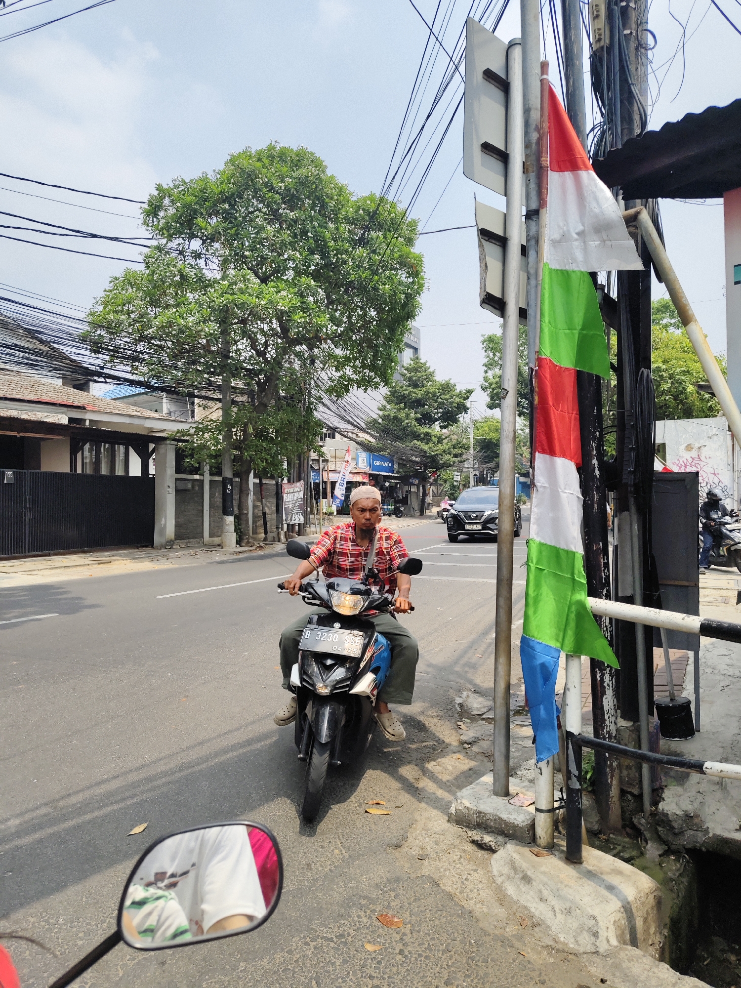 A man wearing a red and white plaid shirt and a white skullcap rides a black motorcycle on a street in South Jakarta, Indonesia. He is looking forward, and his hands are on the handlebars. The motorcycle has a license plate that reads "B 3230 SSE". In the background, there are trees, buildings, and utility poles with many wires. A flag with red and white stripes and green stripes is attached to a pole on the right side of the image. The sky is cloudy. The road is asphalt and appears to be in good condition. There are some fallen leaves on the ground. The image appears to have been taken during the daytime.