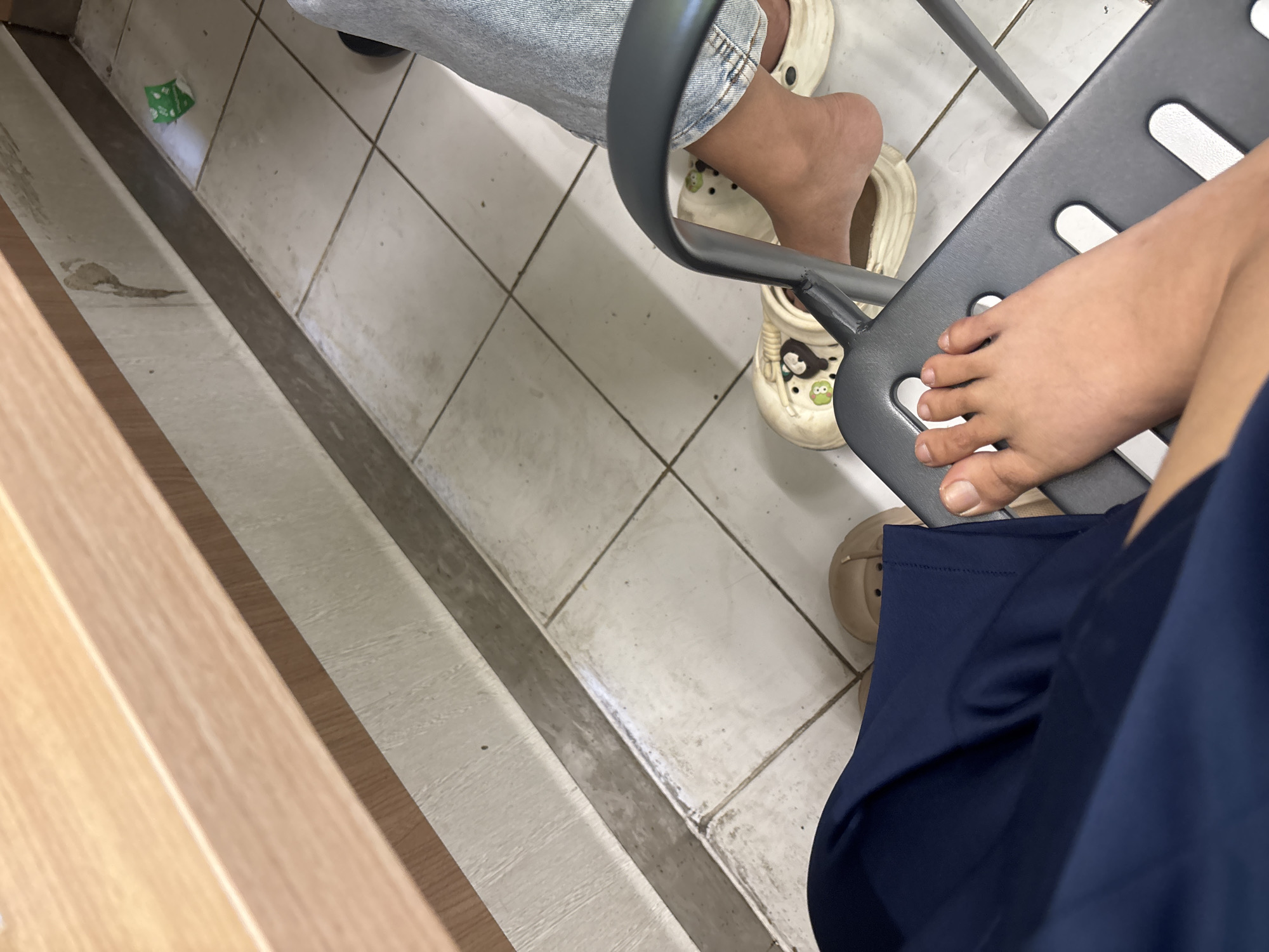 A low-angle, close-up shot captures a person's feet and lower legs in an indoor setting, possibly a waiting area or office in East Jakarta, Indonesia. The floor is tiled with off-white squares, and some dirt is visible in the grout lines. To the left, a dark blue garment, likely pants or a skirt, is draped. In the center and right, two pairs of shoes are visible. One is a light tan slipper, partially obscured by the blue garment. The other is a white Croc-style sandal with cartoon characters printed on it, worn by a person whose legs are clad in light blue jeans. The person in the foreground is resting their bare foot on a gray, perforated chair seat, with their toes clearly visible. A wooden counter or desk runs along the top of the frame, and the wall beneath it is off-white with some staining and peeling. A small, crumpled green piece of paper is on the floor in the background. The lighting appears to be artificial and somewhat dim, casting soft shadows.