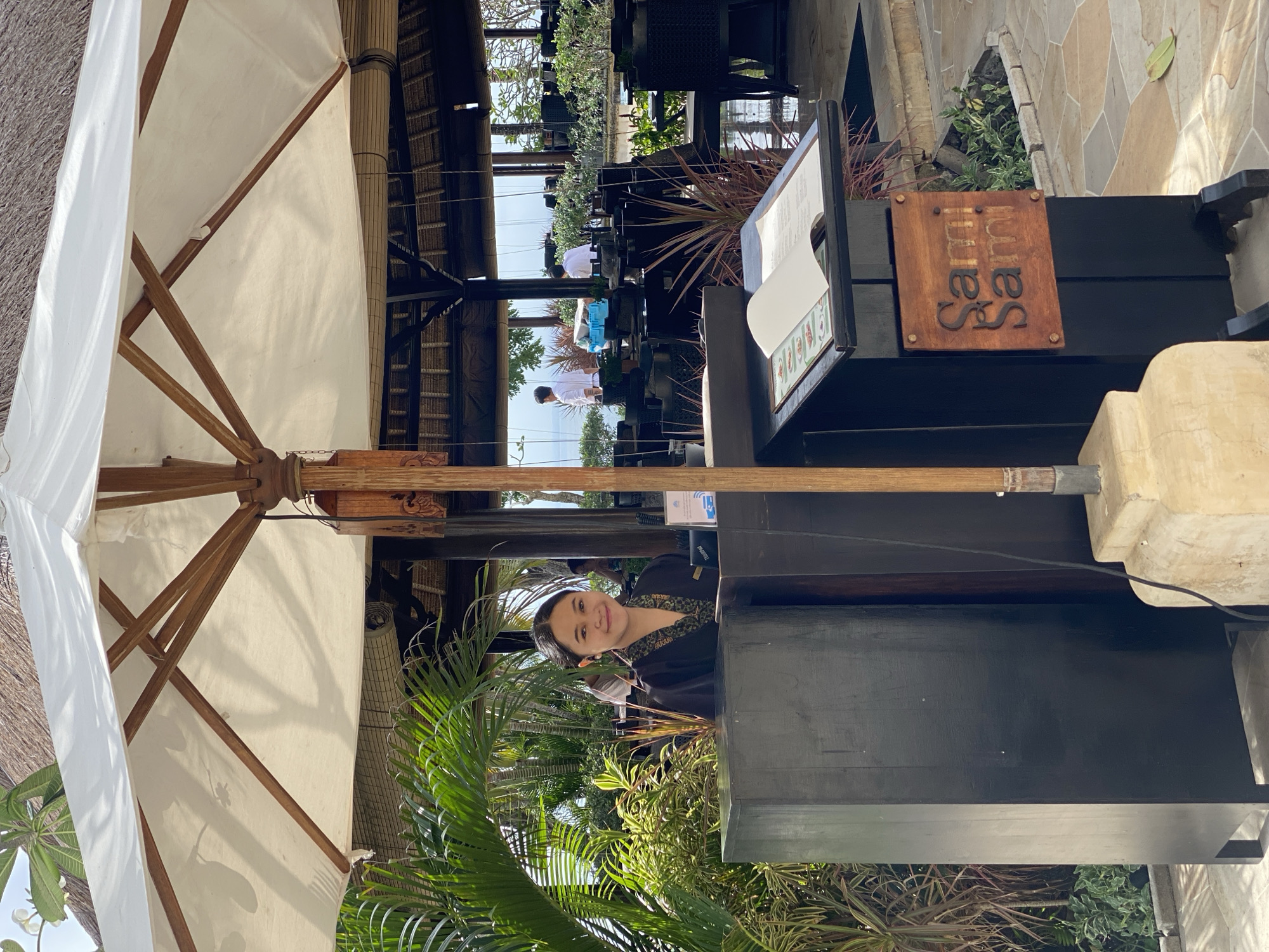 A young woman with dark hair pulled back, wearing a dark uniform with a patterned collar, stands behind a dark wooden counter at the entrance of an outdoor restaurant. She is smiling and looking at the camera. Tropical plants surround the area. An open menu is displayed on a stand in front of her, featuring a wooden sign with the restaurant's name, "Sami Sami." In the background, the restaurant has a thatched roof and outdoor seating. The scene is set in Kuta Selatan, Indonesia.